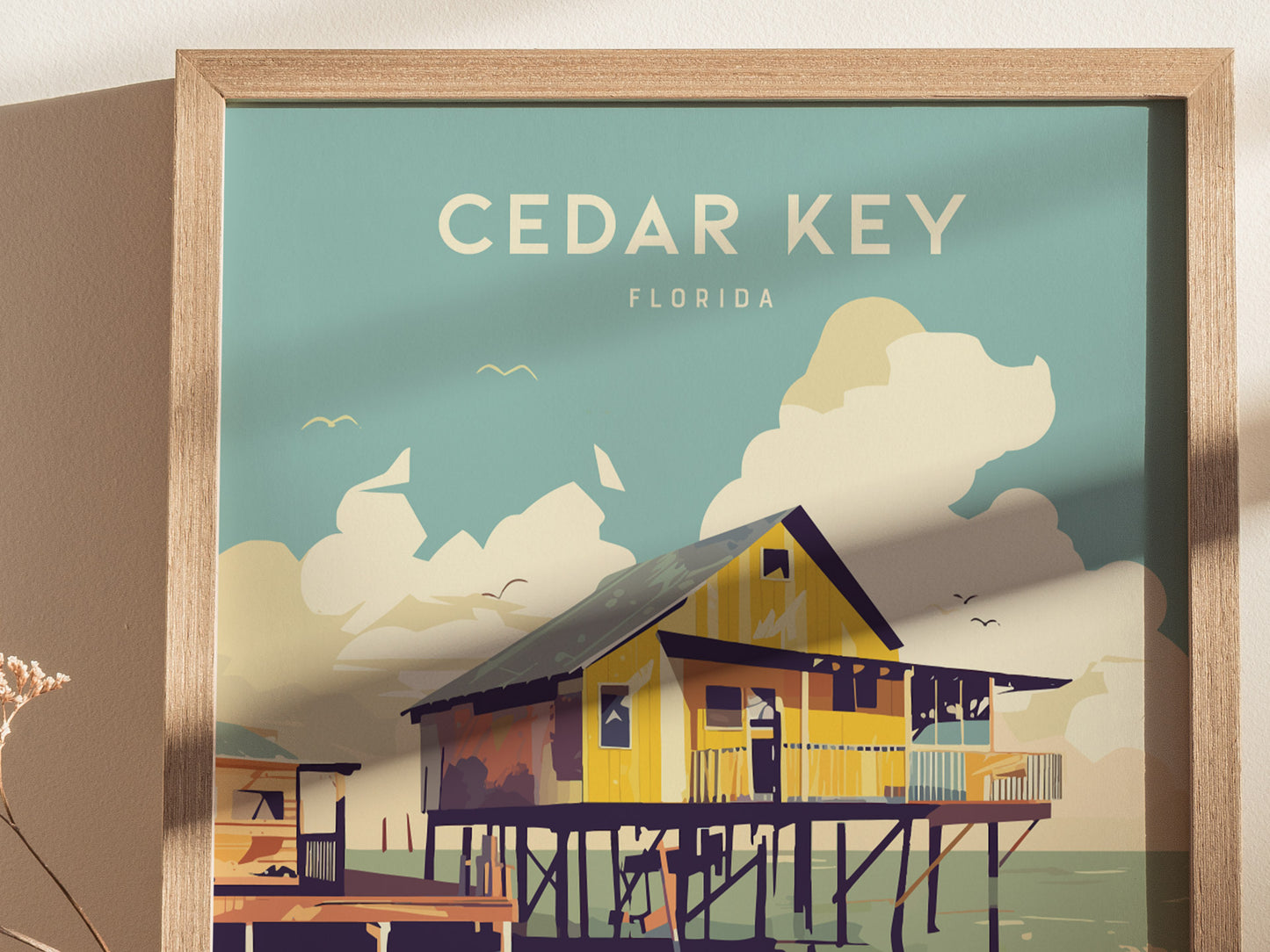 A framed poster depicting a stylized illustration of a stilt house over coastal waters with Cedar Key, Florida text. The background features clouds and birds against a blue sky.