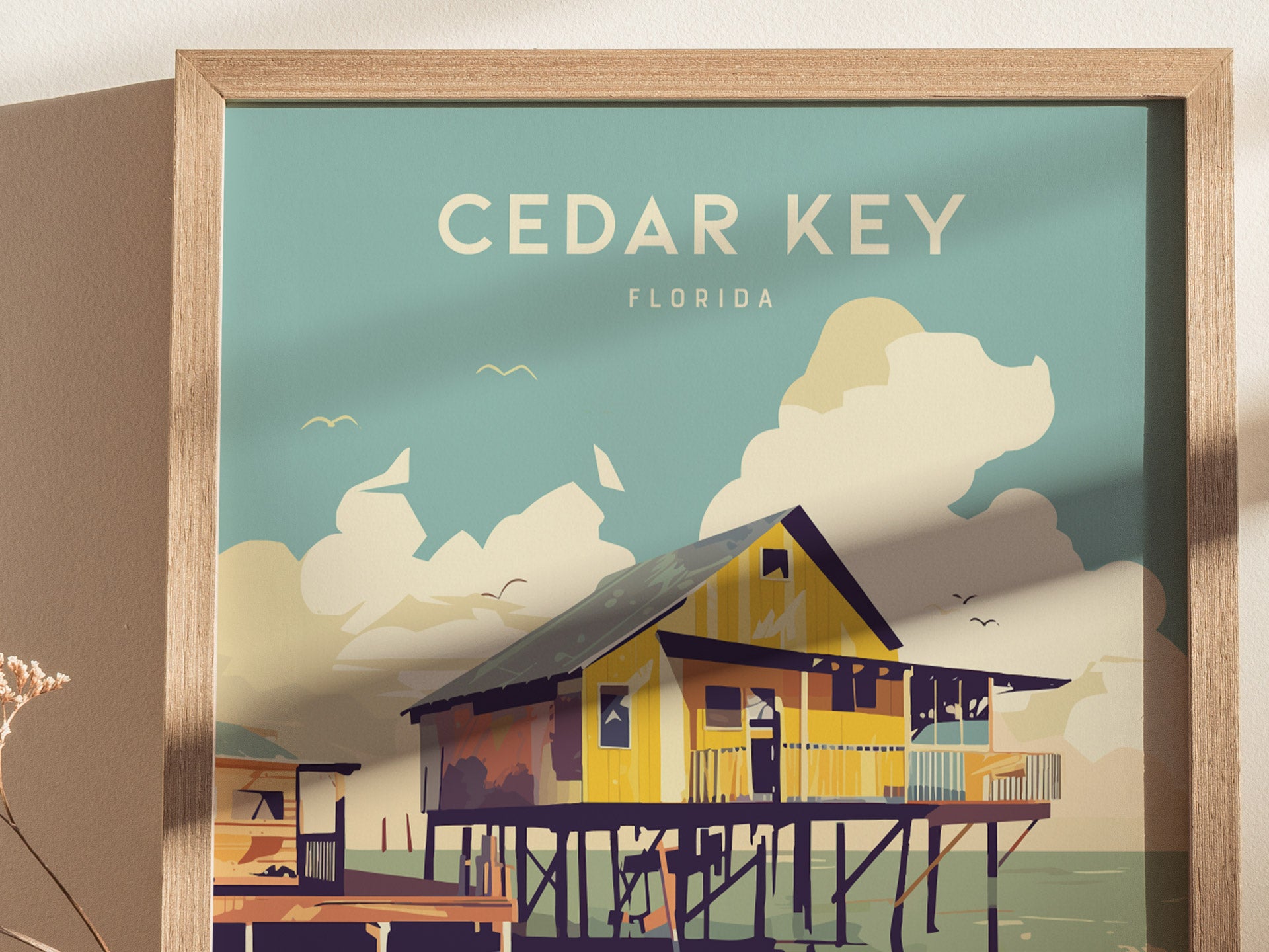 A framed poster depicting a stylized illustration of a stilt house over coastal waters with Cedar Key, Florida text. The background features clouds and birds against a blue sky.