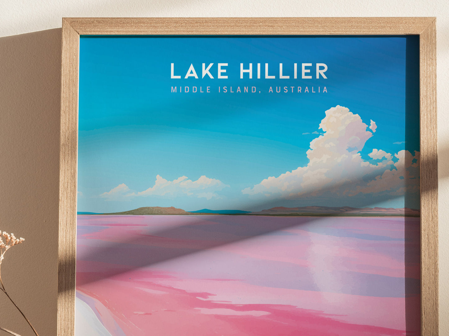 A framed poster of Lake Hillier, featuring vibrant pink water contrasted with a blue sky and fluffy clouds, hangs against a beige wall.