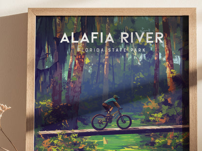 A framed poster of Alafia River State Park shows a mountain biker crossing a wooden bridge over a lush, green landscape with tall trees and vibrant foliage.