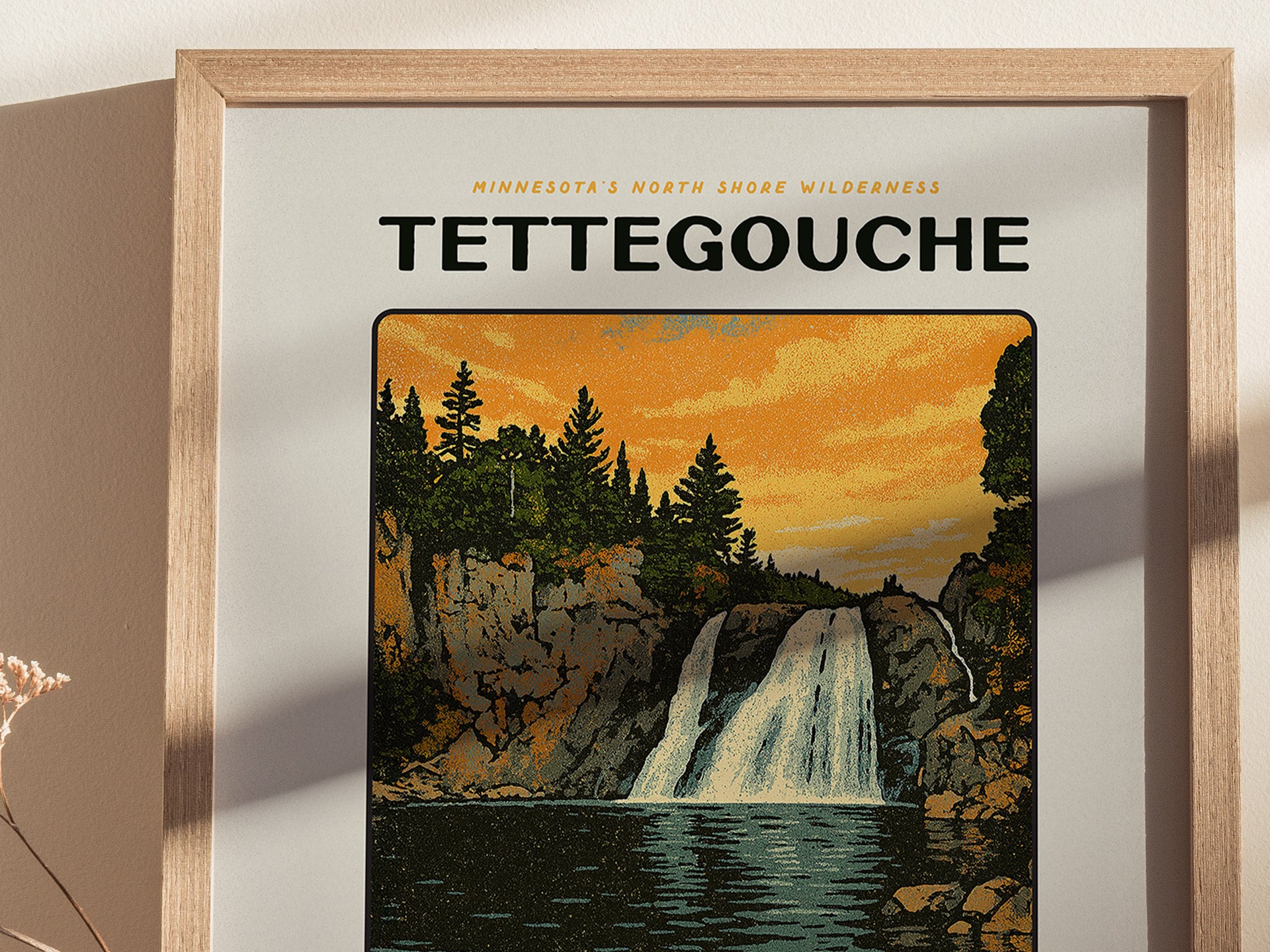 A framed poster depicts a waterfall cascading between rocky cliffs under a vibrant orange sky, with pine trees lining the top. The text reads Minnesotas North Shore Wilderness Tettegouche.