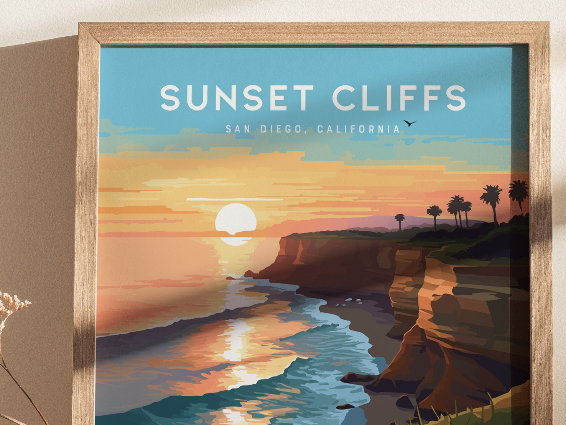 Framed artwork depicting a vibrant sunset over cliffs and ocean waves, with palm trees silhouetted against the colorful sky. Text reads Sunset Cliffs, San Diego, California.