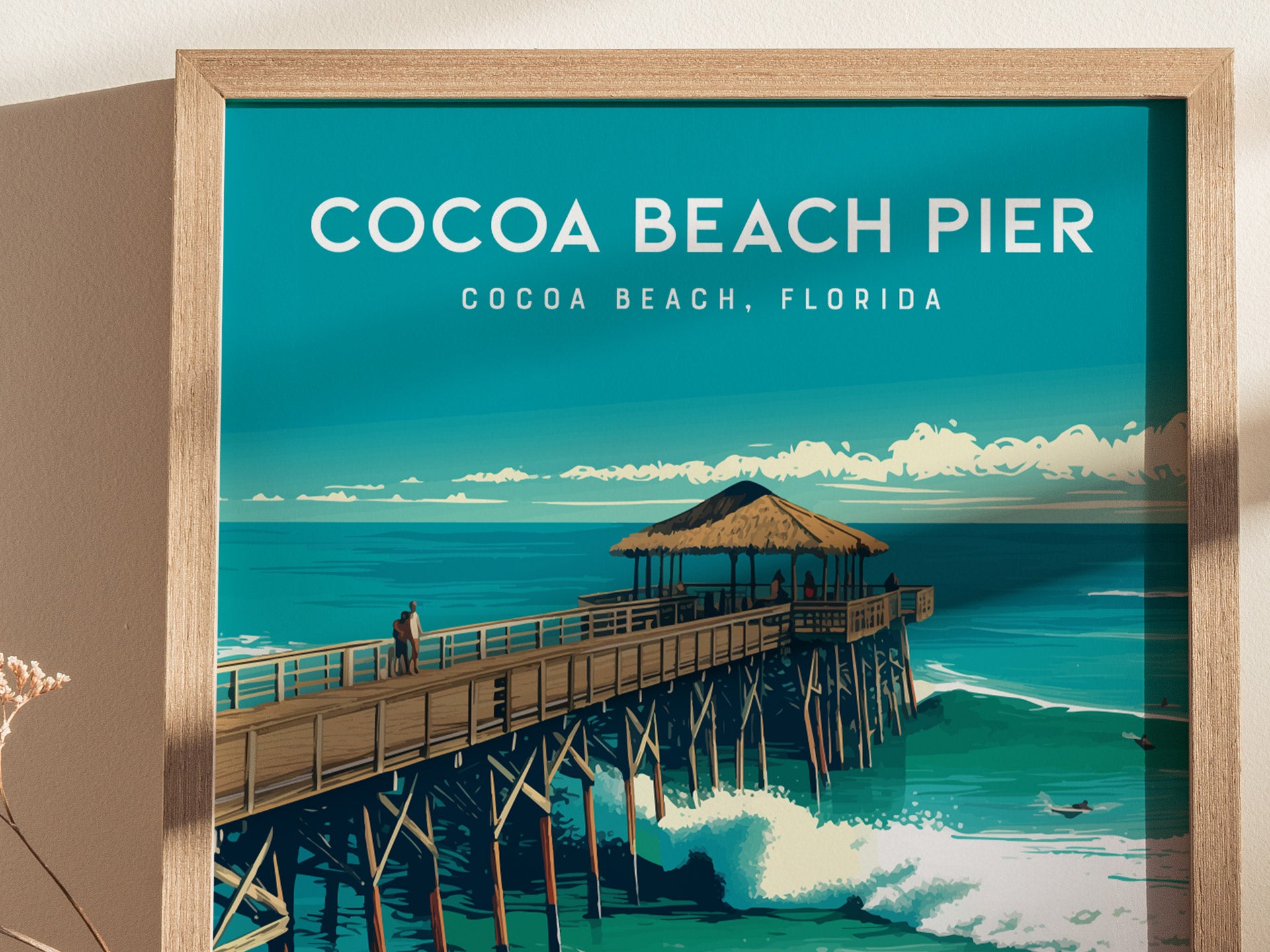 A framed poster featuring the Cocoa Beach Pier, with a thatched-roof pavilion over the ocean and surfers riding waves below, set against a vibrant blue sky.