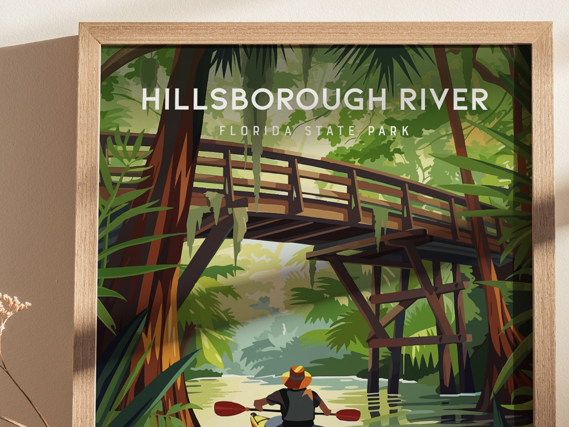 A framed poster depicts a kayaker paddling beneath a rustic wooden bridge in a lush, green forest. The scene is filled with towering trees and hanging Spanish moss.