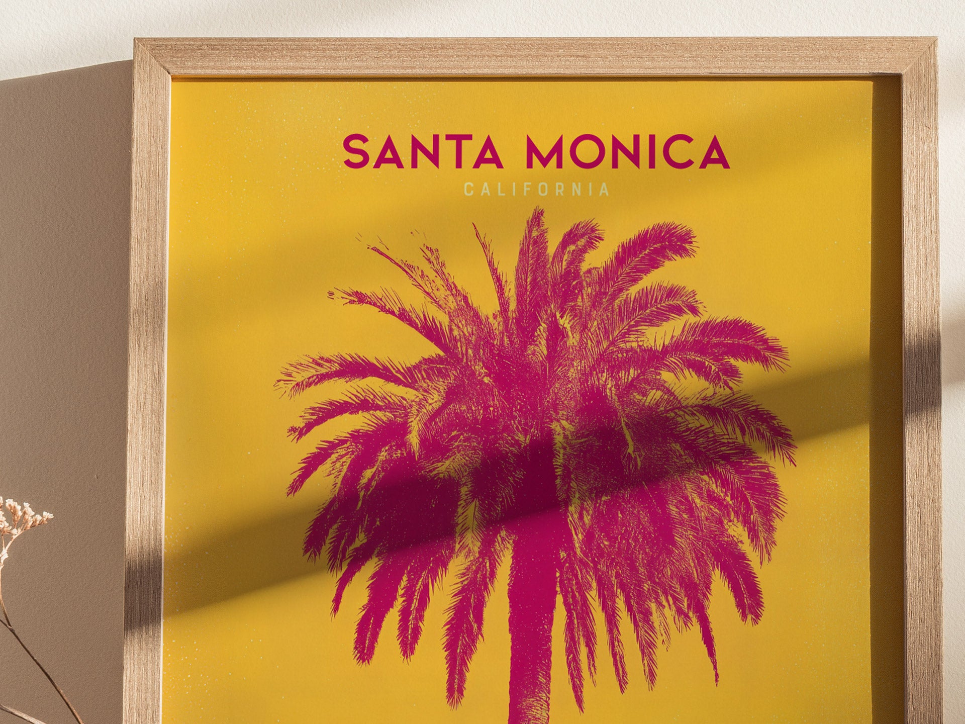A framed poster features a vibrant magenta palm tree against a bright yellow background with the words Santa Monica California at the top. A shadow falls diagonally across the image.
