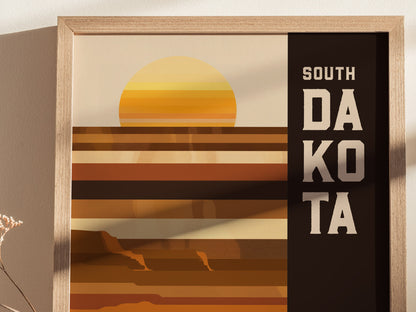 A framed poster features a geometric design with earthy tones and a stylized sun, alongside bold text reading South Dakota. Shadows and a dried flower are visible on the left.