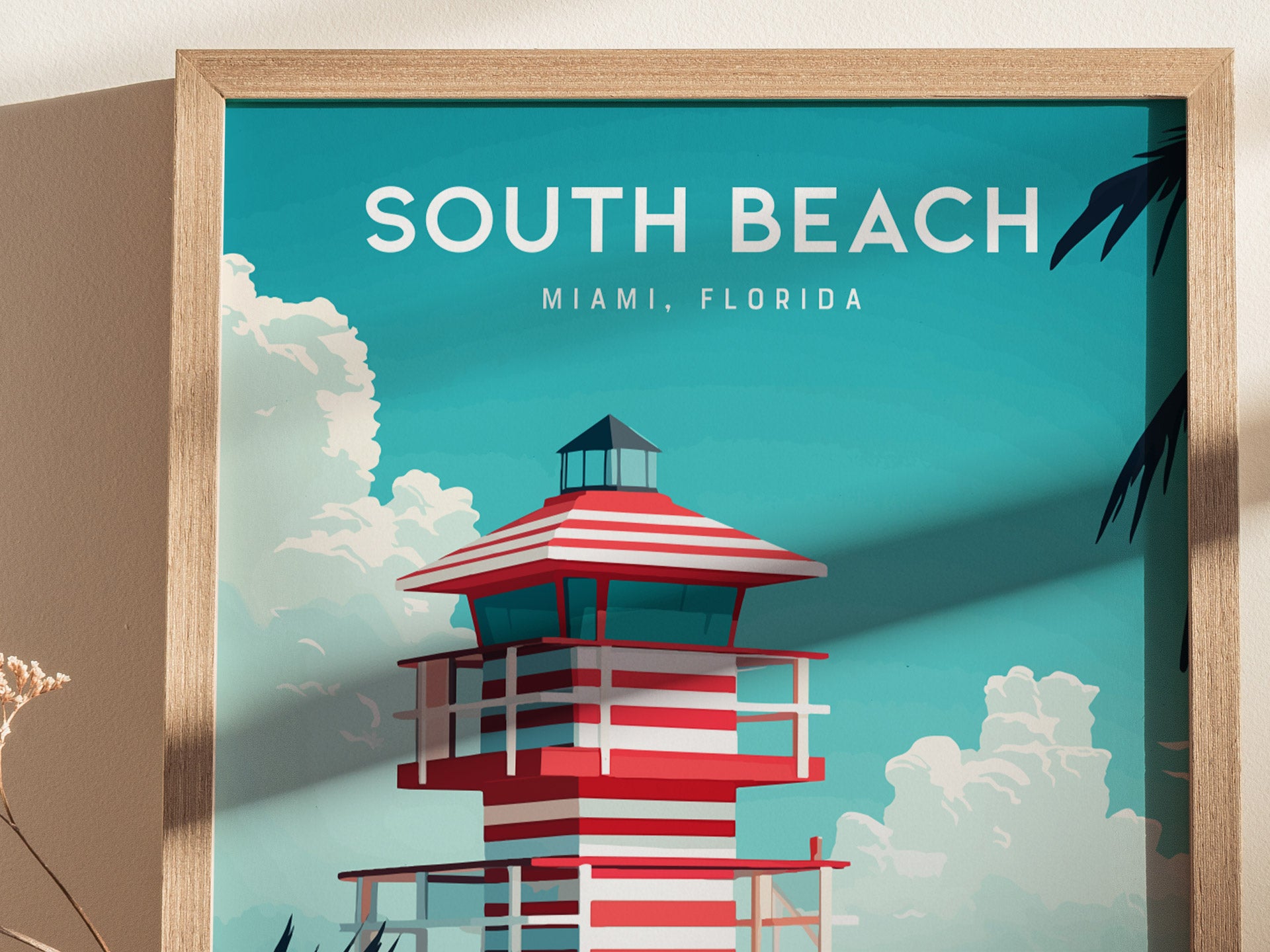 A framed poster depicts a stylized lifeguard tower with red and white stripes against a bright blue sky, featuring clouds and palm leaves. The words South Beach, Miami, Florida are prominently displayed at the top.