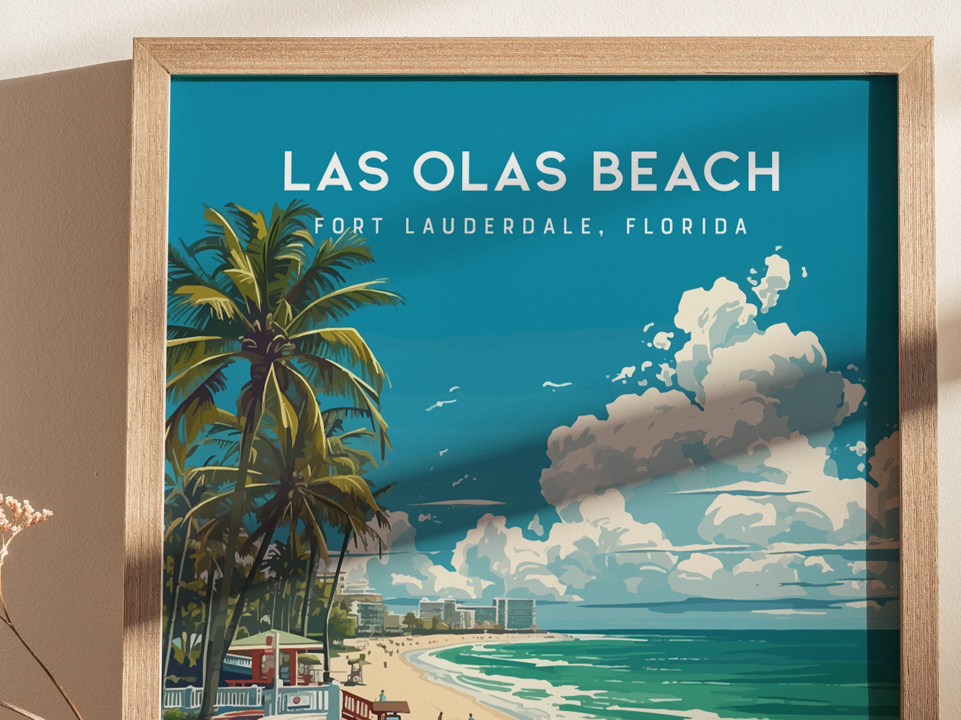 A framed Las Olas Beach poster features palm trees, a lifeguard stand, and a beach scene with clouds in Fort Lauderdale, Florida. The artwork has a vibrant, modern design.