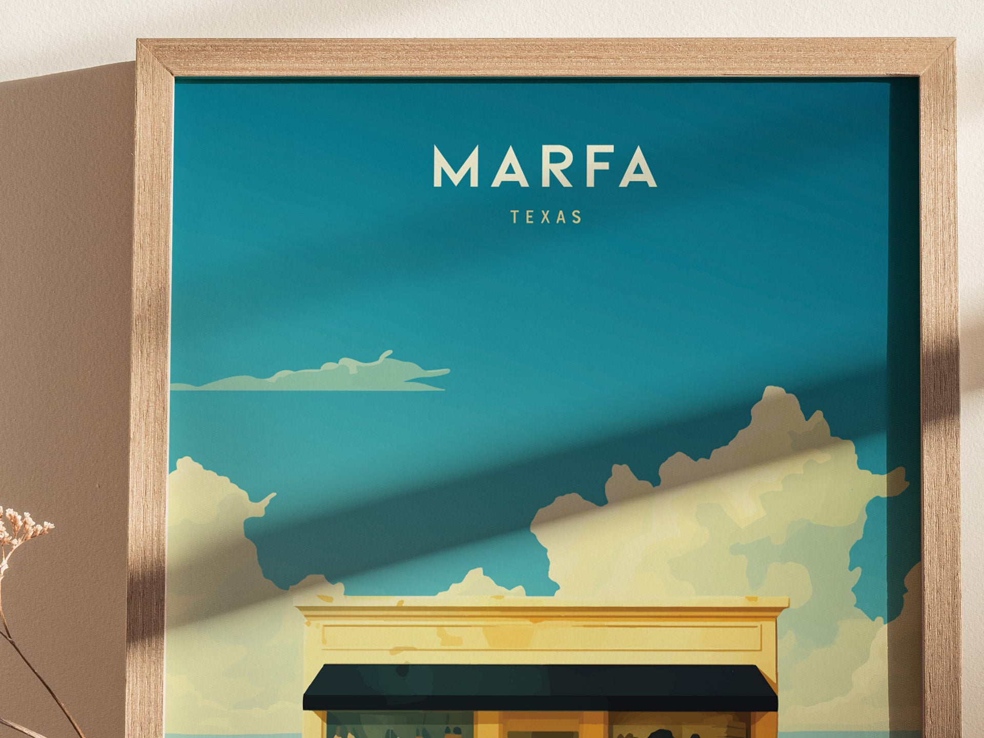 A framed poster features a stylized illustration of a building under a clear blue sky with fluffy clouds, labeled Marfa Texas. Sunlight casts a shadow on the artwork.