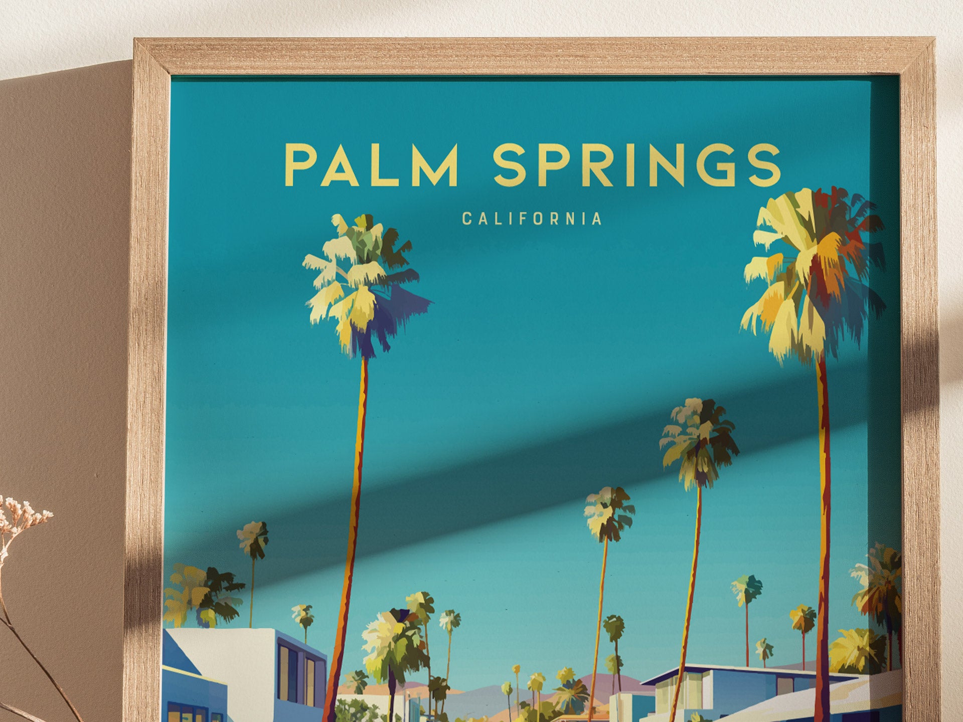 A framed poster with Palm Springs California text showcases stylized palm trees against a vibrant blue sky, with modern buildings in the foreground, capturing a mid-century desert aesthetic.
