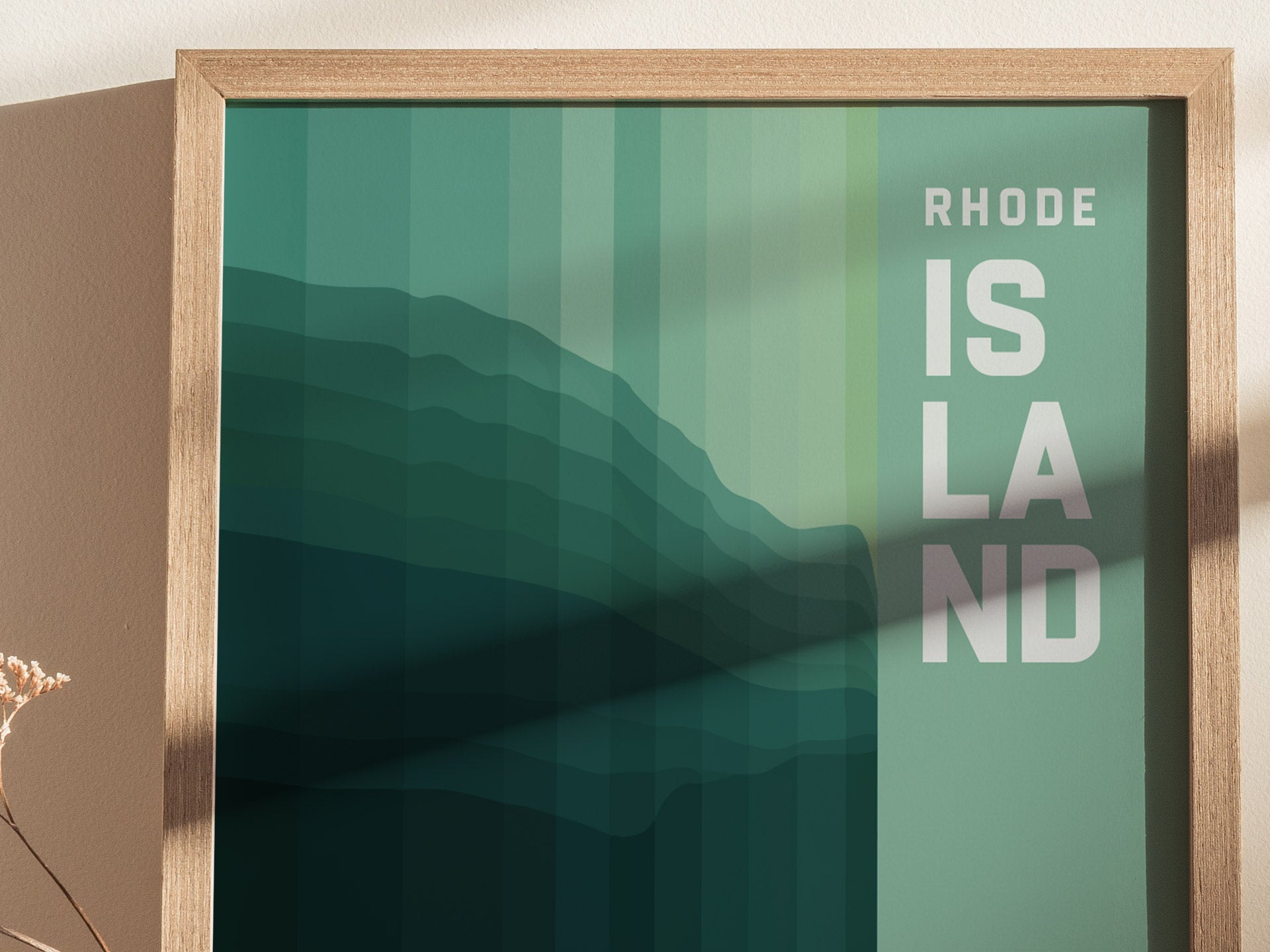 Framed abstract poster featuring layered green shapes with the text RHODE ISLAND in bold white letters. The artwork is displayed against a neutral wall with a dried flower nearby.