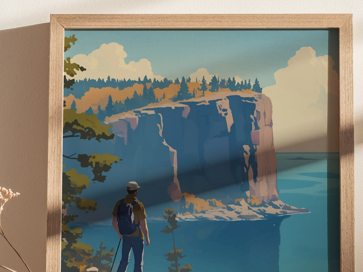 A framed poster depicting a hiker with a backpack gazing at dramatic cliffs beside a calm blue lake under a partly cloudy sky. The scene is rich with autumnal colors.