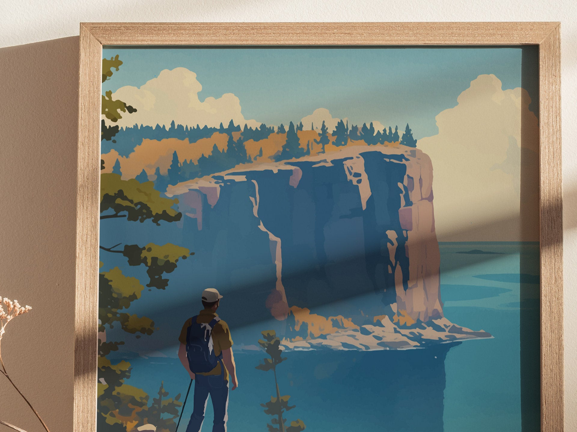 A framed poster depicting a hiker with a backpack gazing at dramatic cliffs beside a calm blue lake under a partly cloudy sky. The scene is rich with autumnal colors.