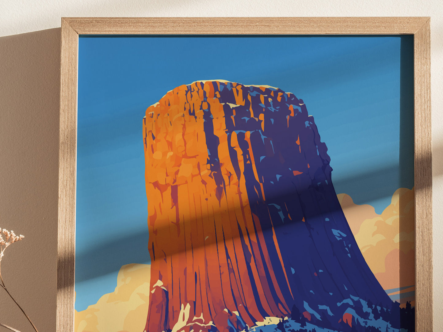 A framed poster of Devils Tower, featuring vibrant orange and blue hues against a clear blue sky, is displayed on a wall. Shadows add depth to the artwork.