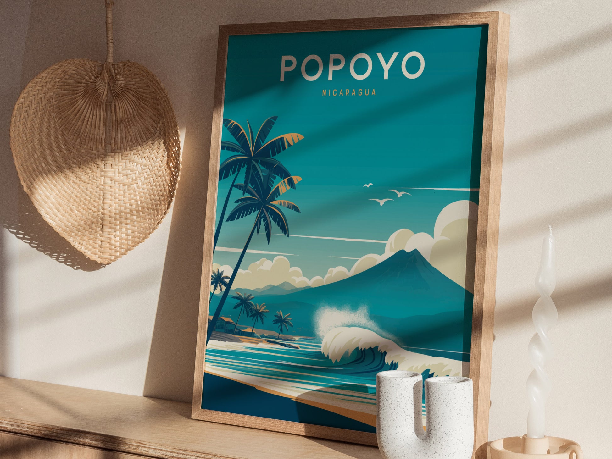 A framed Popoyo Nicaragua surf art poster features palm trees, waves, and a mountain under a blue sky. Its placed on a shelf with a woven fan and decorative candle.