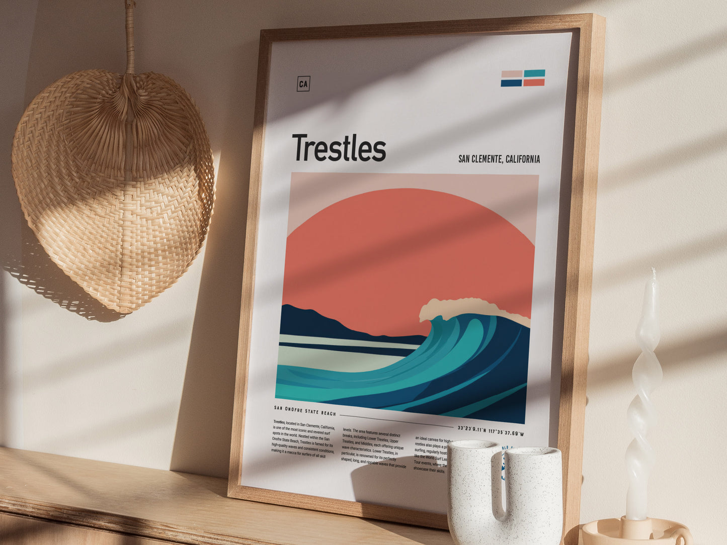 A framed Trestles surf poster with minimalist wave and sunset design sits on a wooden shelf, accompanied by a woven wall decoration and a modern candle.