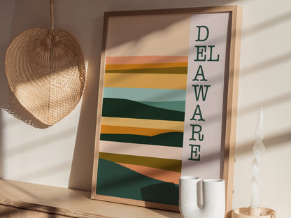 A framed minimalist poster featuring abstract, layered shapes in earthy tones with the word Delaware vertically aligned. It is displayed on a wooden surface next to a woven decor piece.