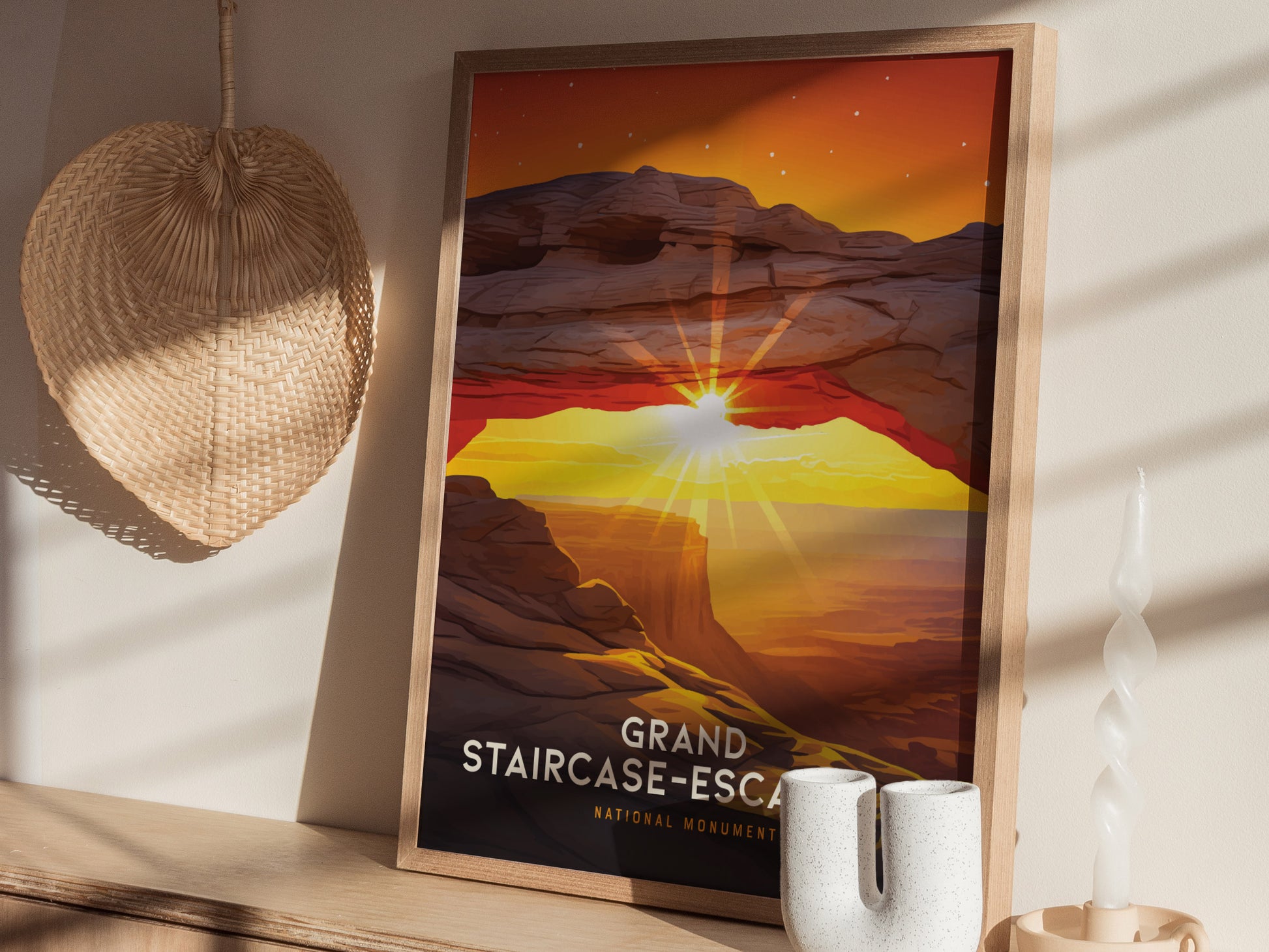 A framed poster of Grand Staircase–Escalante features a sunset arch with vibrant red and yellow hues. The artwork rests on a shelf beside a woven fan and decorative candle.