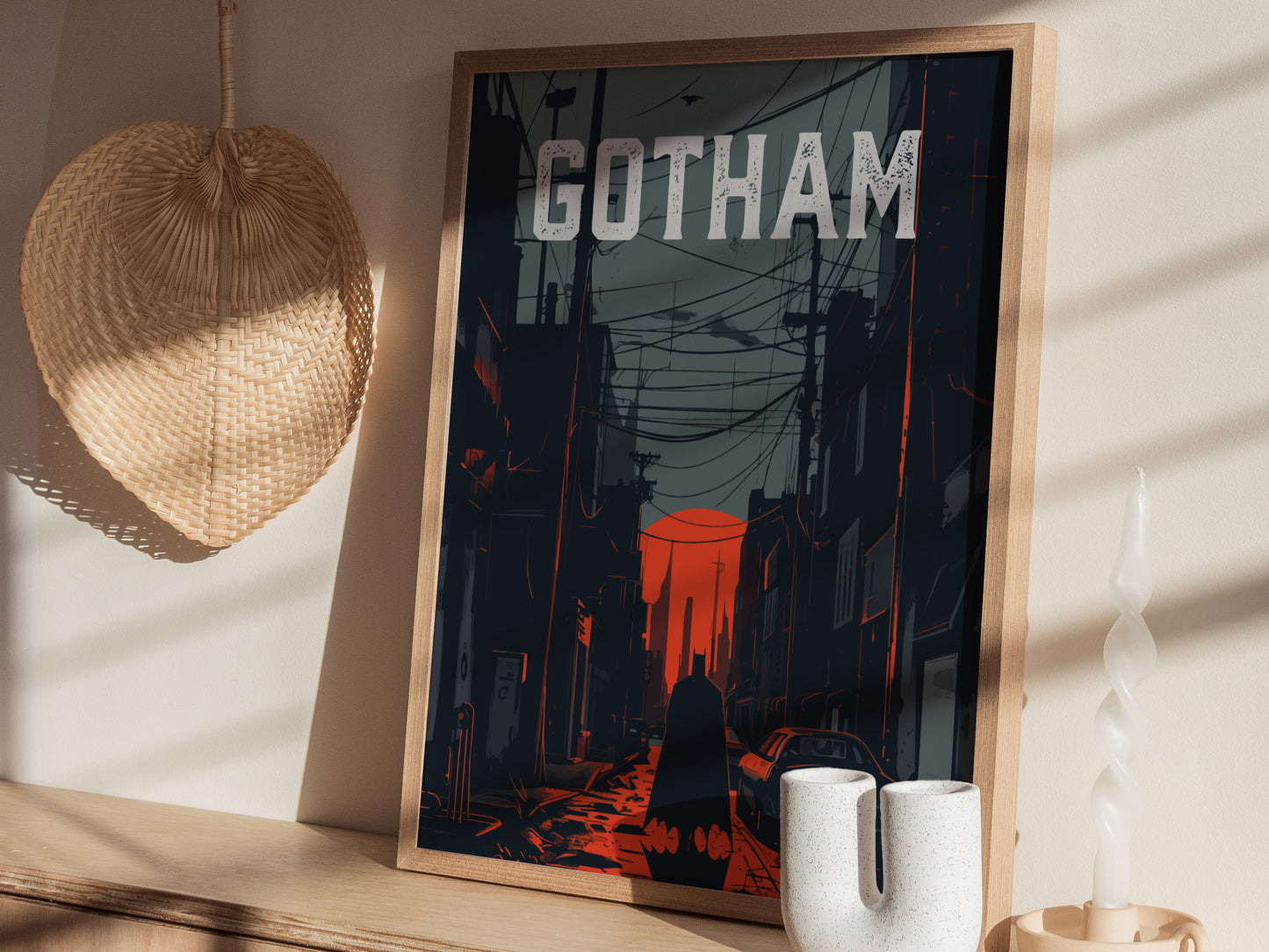A framed poster titled GOTHAM depicts a noir-style cityscape with dark buildings and red highlights. Its displayed on a wooden shelf next to decorative items, including a woven fan and a twisted candle.