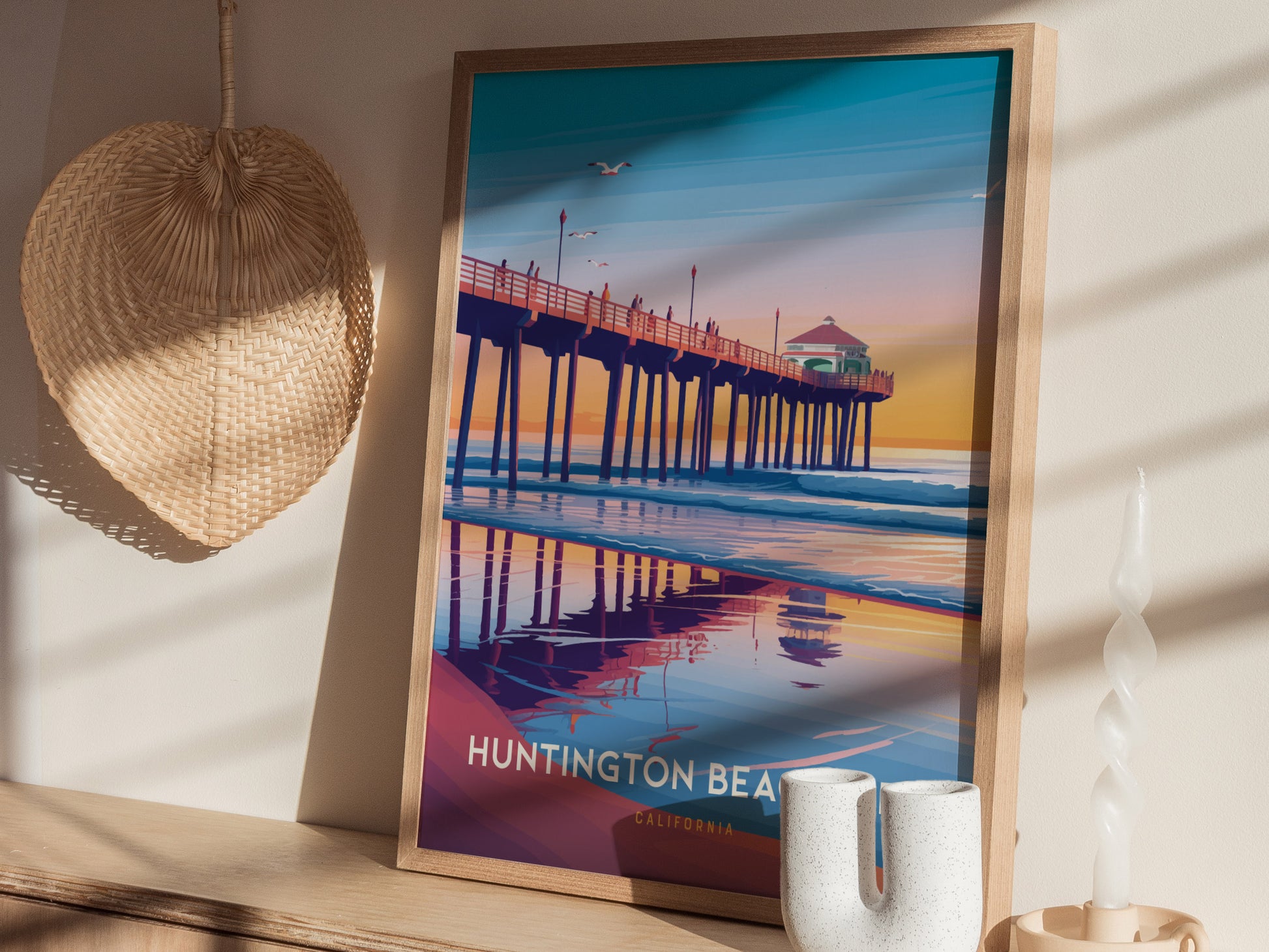 A framed poster of Huntington Beach Pier at sunset, featuring vibrant colors and reflections, rests on a wooden surface beside a woven hat and twisted candle.