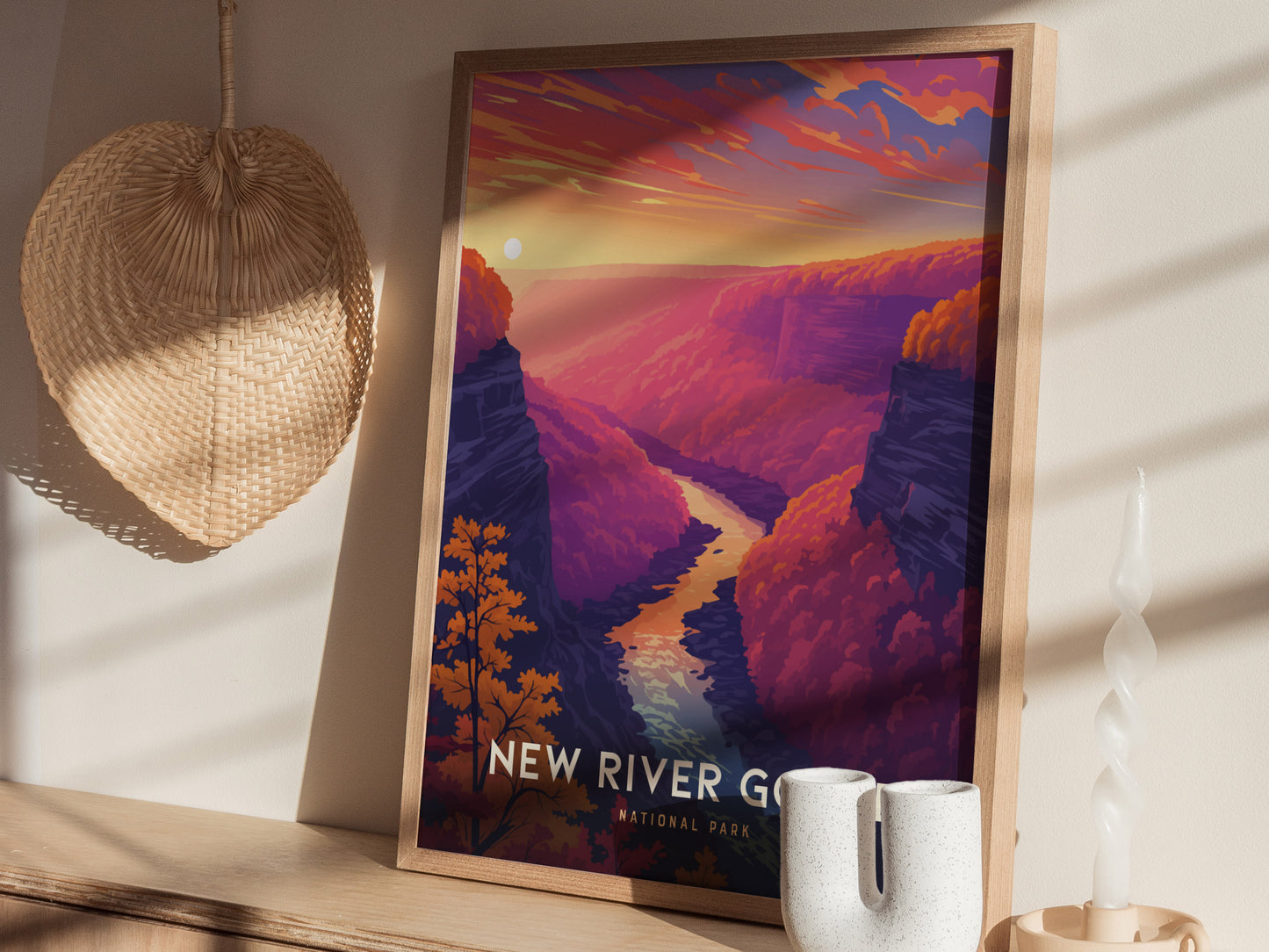 A framed poster of New River Gorge, featuring a colorful landscape with a winding river, sits on a shelf next to a wicker fan, candle, and ceramic vase.
