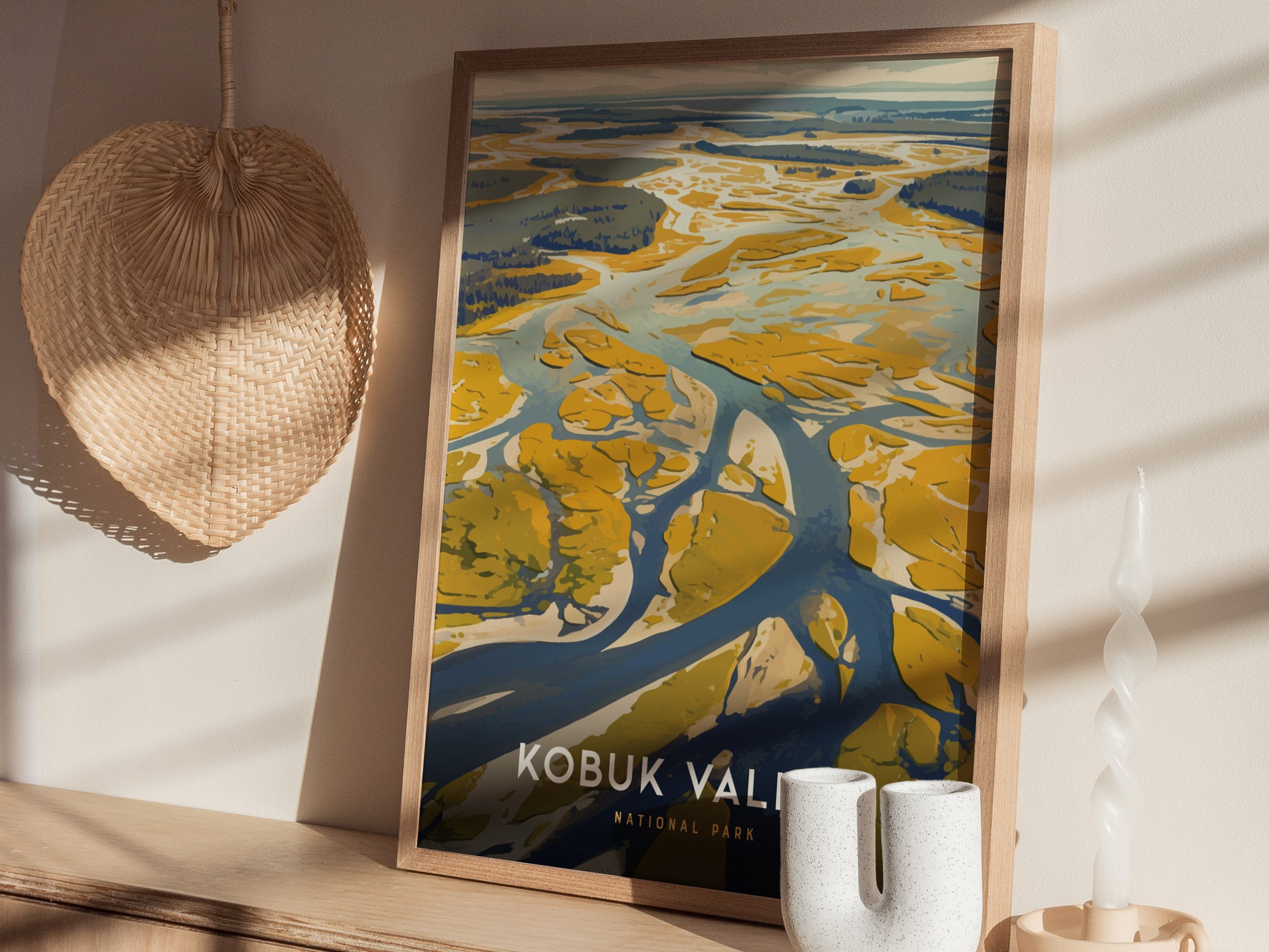 A framed poster of Kobuk Valley National Park with abstract sand dunes and rivers sits on a wooden shelf, accompanied by a woven basket and a decorative candle.