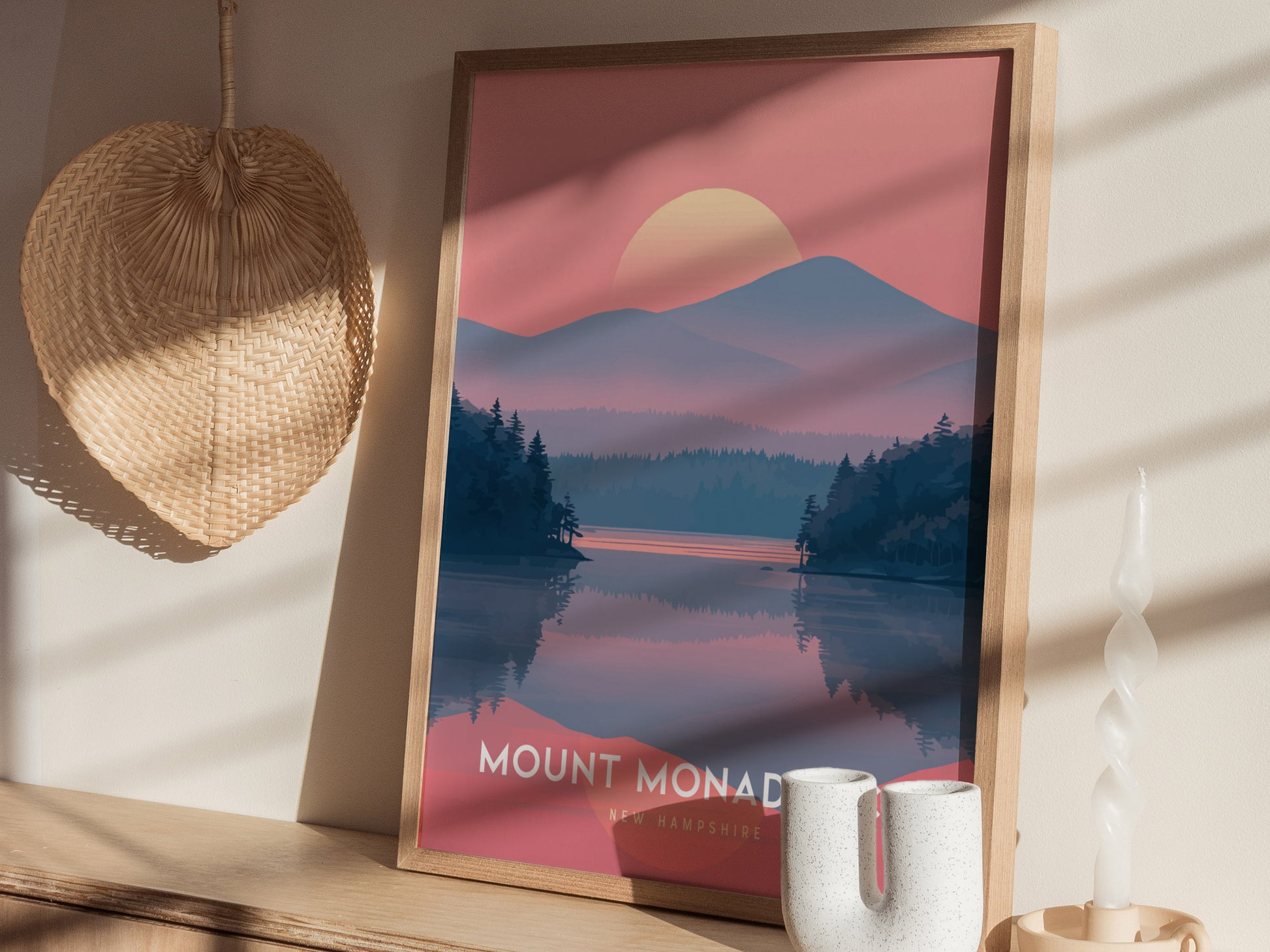 A framed Mount Monadnock poster with a sunset landscape sits on a wooden shelf beside a woven wall hanging and a white ceramic candle holder.