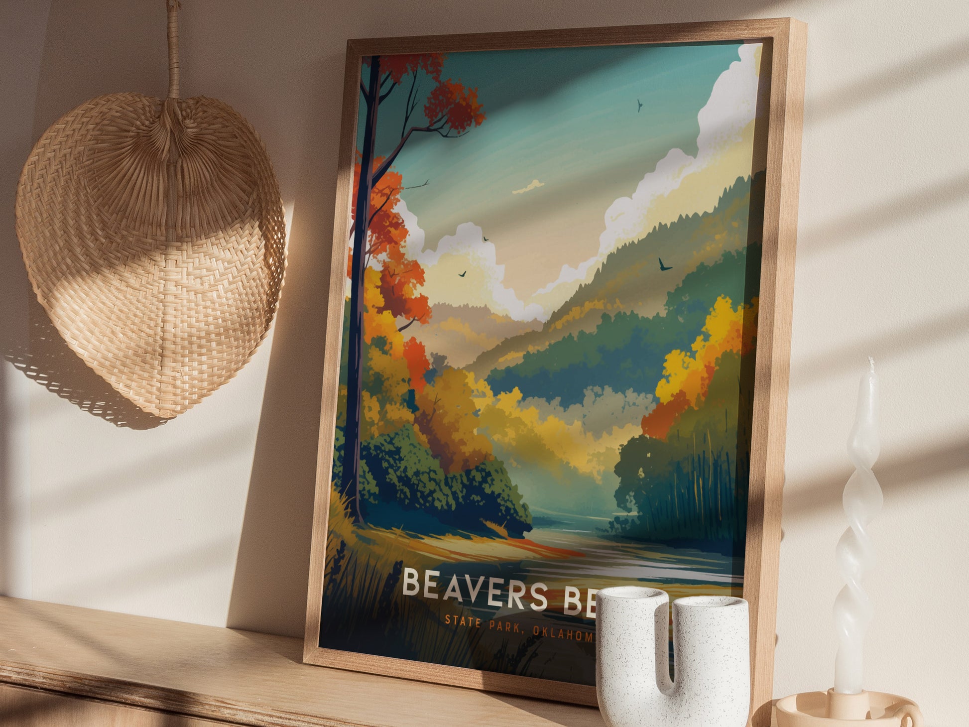 A framed poster of Beavers Bend State Park with colorful trees and mountains sits on a wooden shelf, accompanied by a wicker wall decoration and a unique ceramic vase.