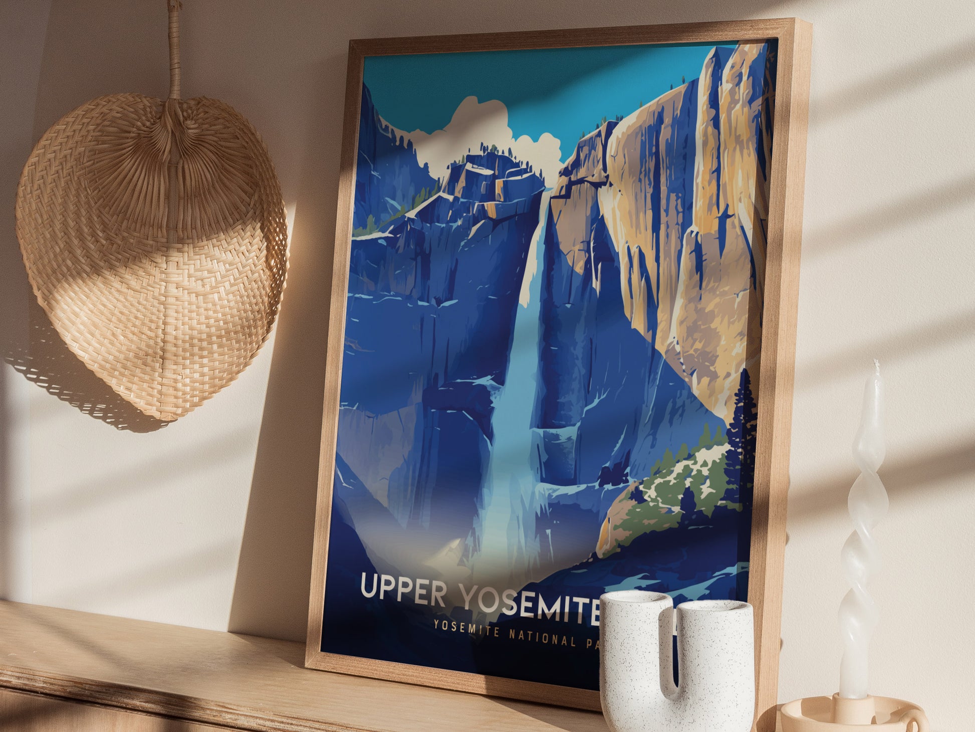 A framed poster of Upper Yosemite Falls features a stylized depiction of the waterfall against granite cliffs. Its displayed on a wooden surface with a woven fan and a twisted.