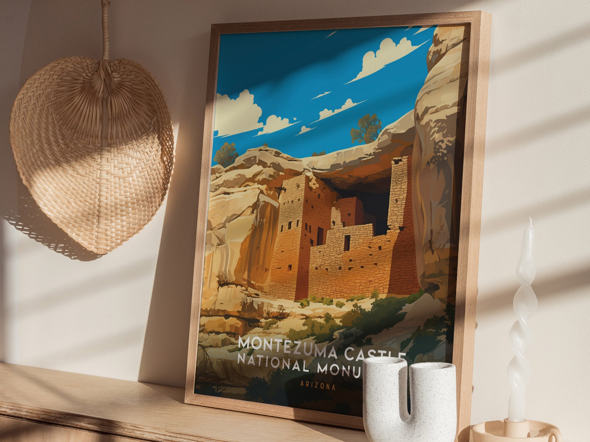 A framed poster of Montezuma Castle National Monument rests on a wooden shelf, next to a woven wall decoration and a twisted white candle. Sunlight casts shadows across the scene.