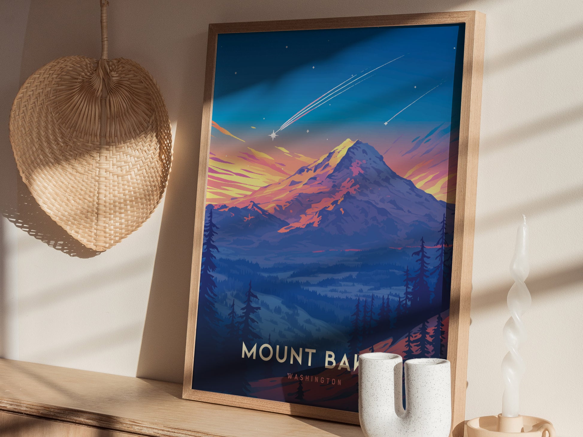 A framed Mount Baker poster sits on a shelf, showcasing a colorful sunset over the mountain. Its surrounded by a woven basket and a uniquely shaped white candle.