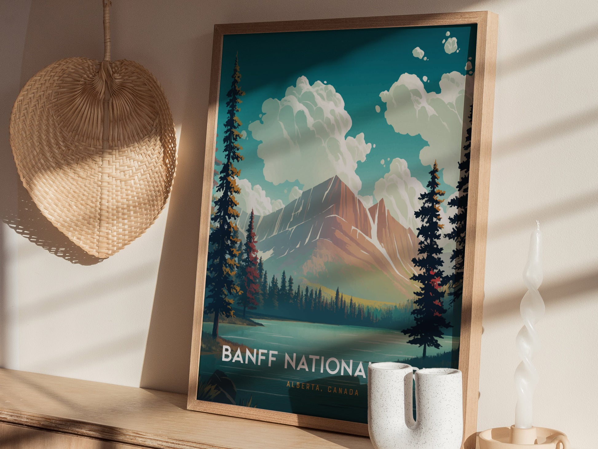 A framed Banff National Park poster shows a vibrant landscape with mountains, trees, and clouds. It sits on a wooden shelf beside a woven basket and a twisted candle.