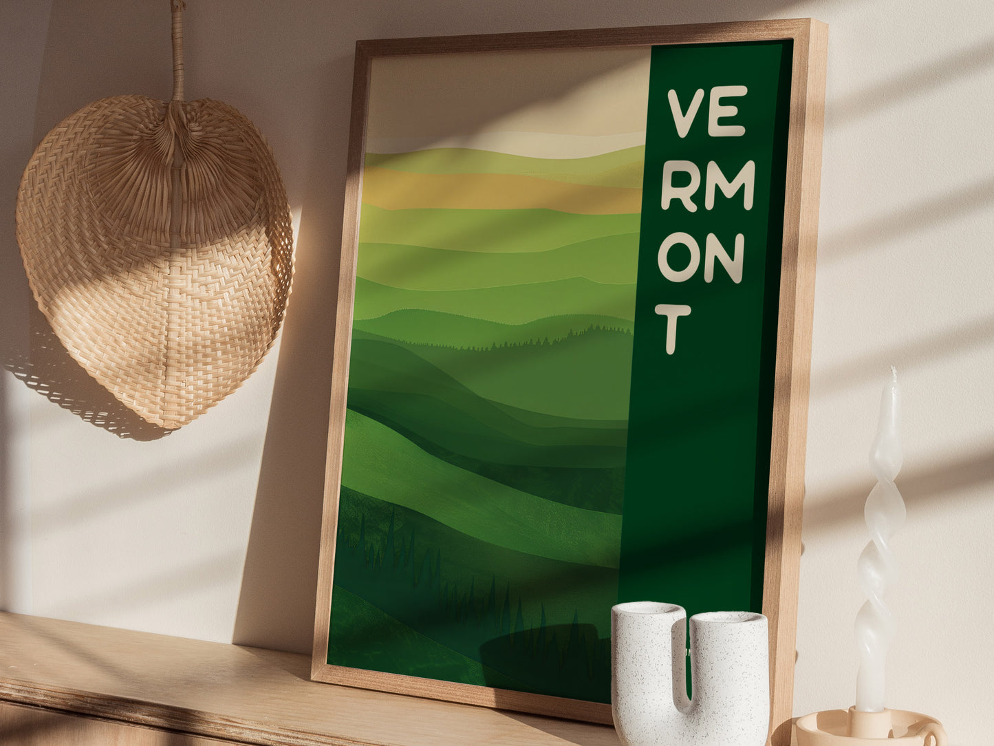 A framed poster with Vermont text features abstract green rolling hills, set on a wooden shelf with a woven wall hanging and decorative ceramic items.