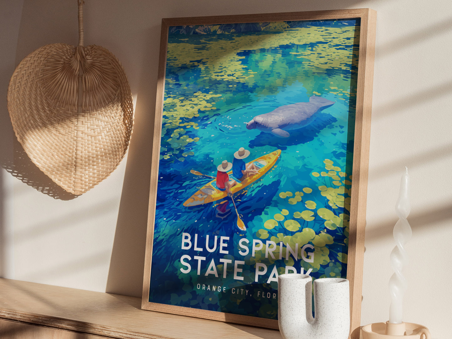 A framed Blue Spring State Park poster features kayakers and a manatee in vibrant blue waters, placed on a shelf with a woven fan and decorative items nearby.