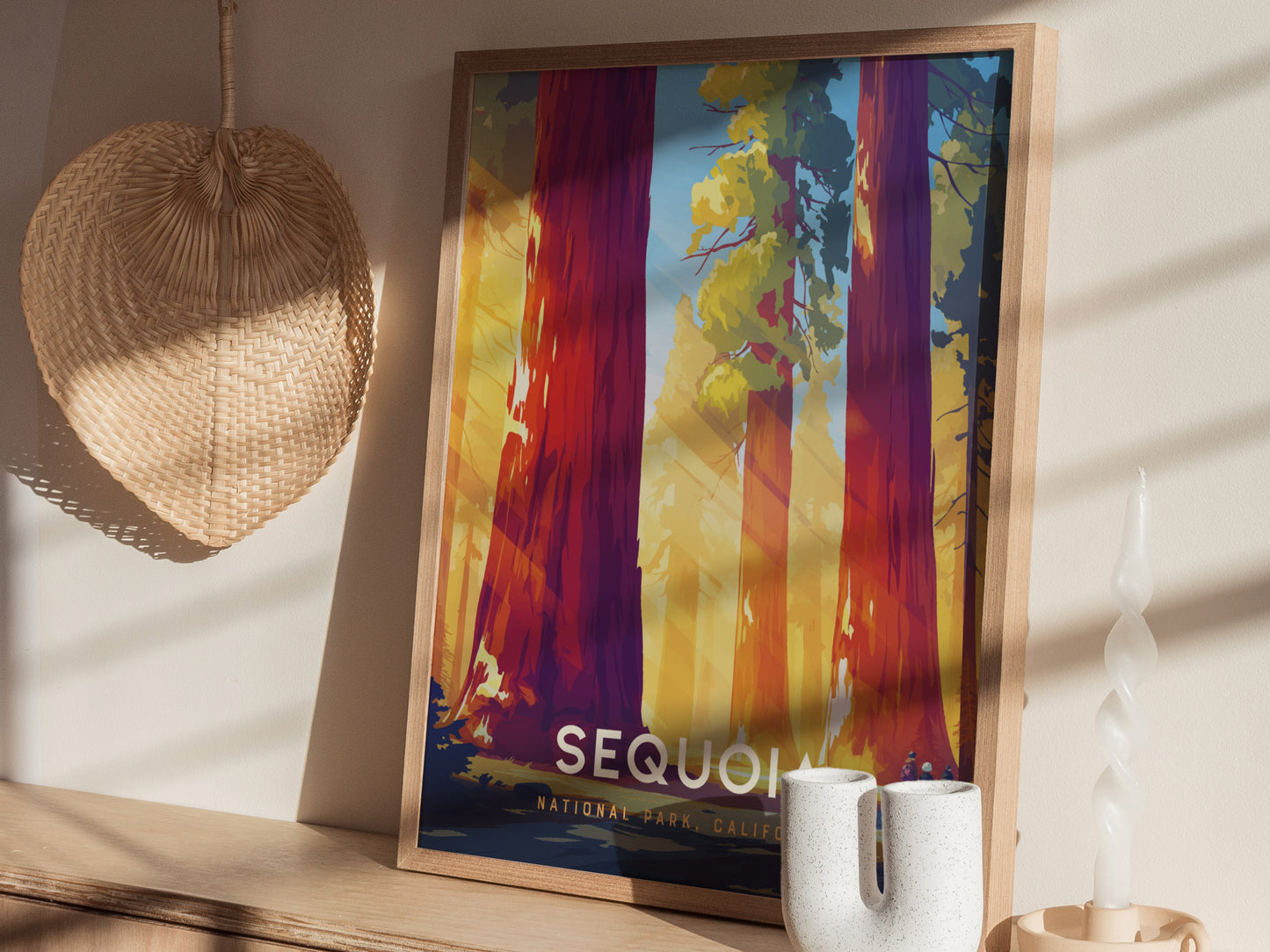 Framed artwork depicting colorful, towering sequoia trees under sunlight is displayed on a wooden shelf, accompanied by a woven basket, a twisted candle, and a white ceramic vase.