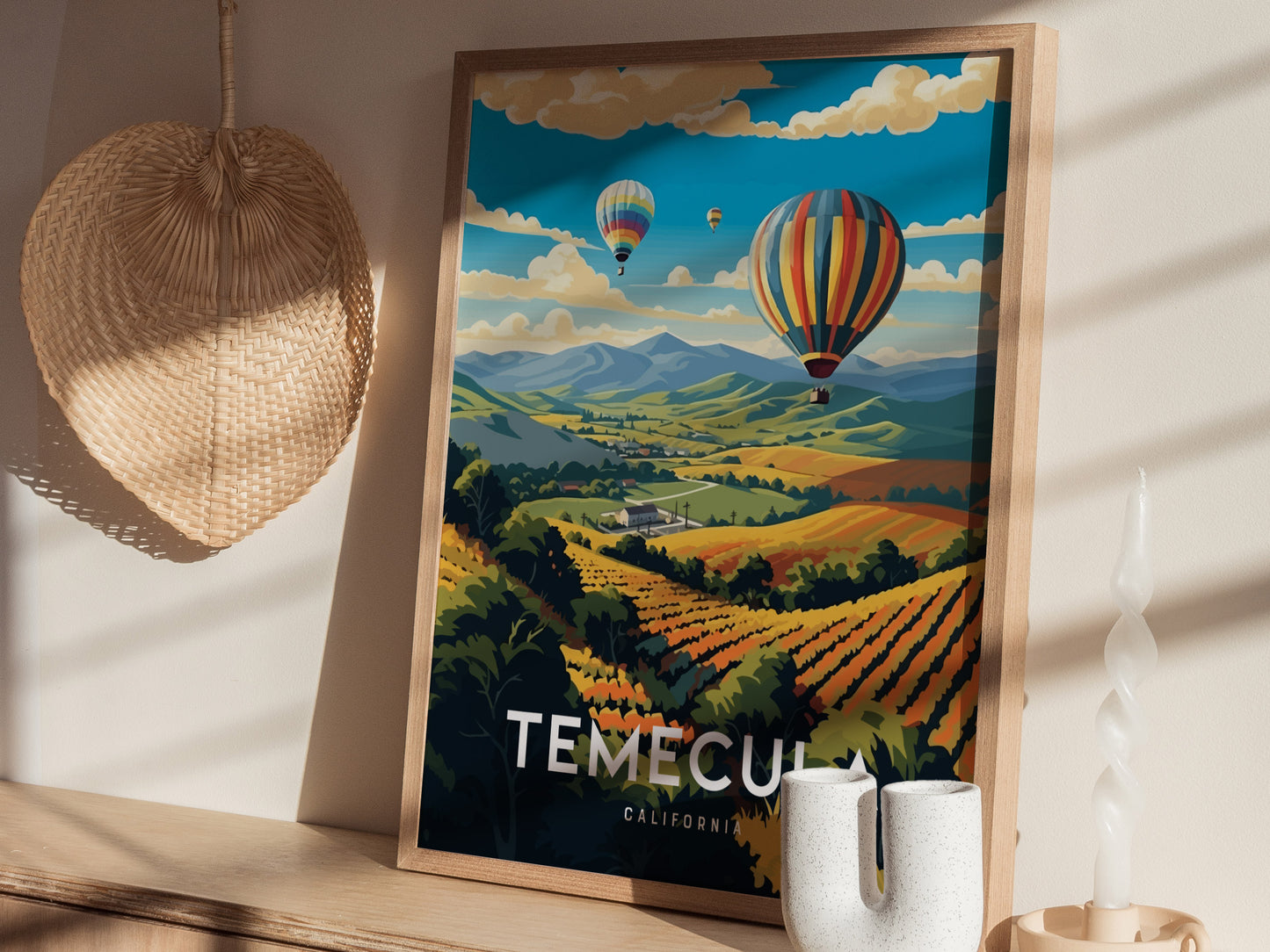 A framed poster of Temecula, California, features vibrant hot air balloons floating over lush vineyards, set against a backdrop of rolling hills. A wicker basket and candle sit nearby.