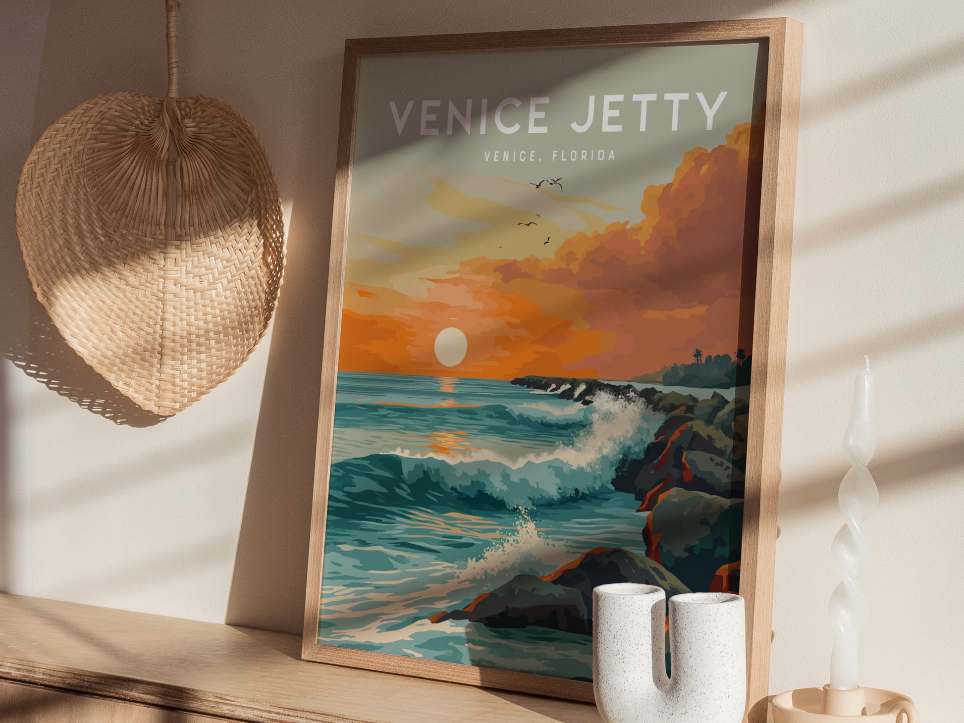 A framed poster titled Venice Jetty depicts a vibrant sunset over ocean waves crashing against a rock jetty. Its displayed on a wooden shelf with a woven wall decor and.