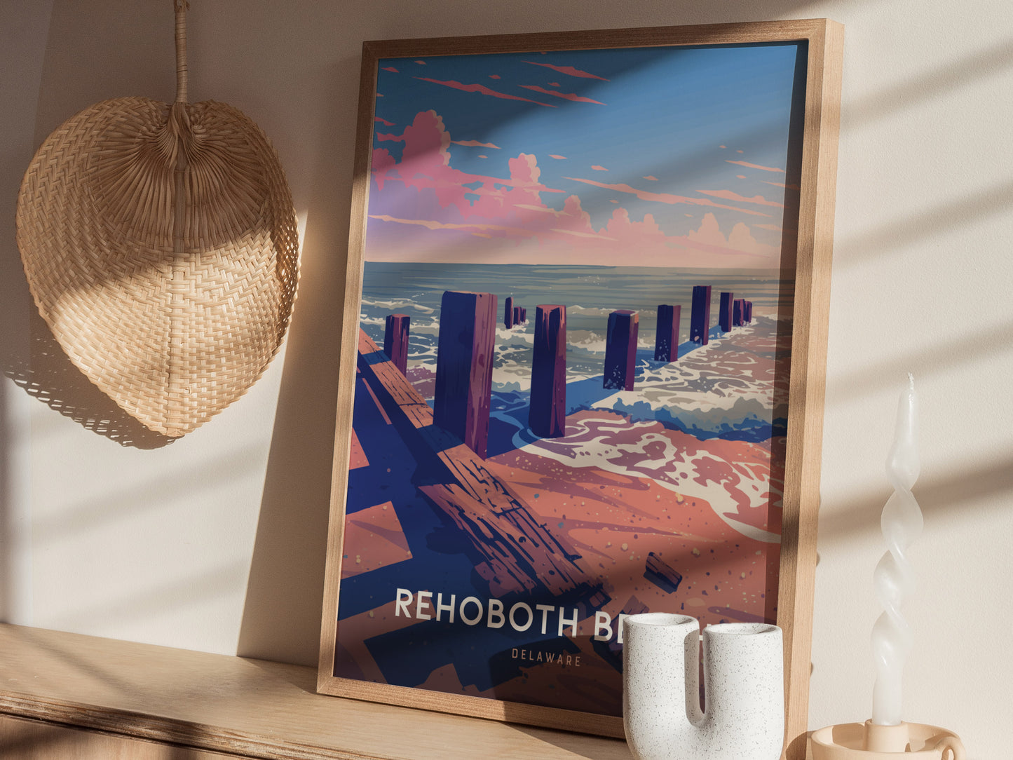 A framed Rehoboth Beach poster depicting a coastal scene with wooden pilings, sandy beach, and ocean waves is displayed on a shelf. A woven hat and decorative candle are nearby.