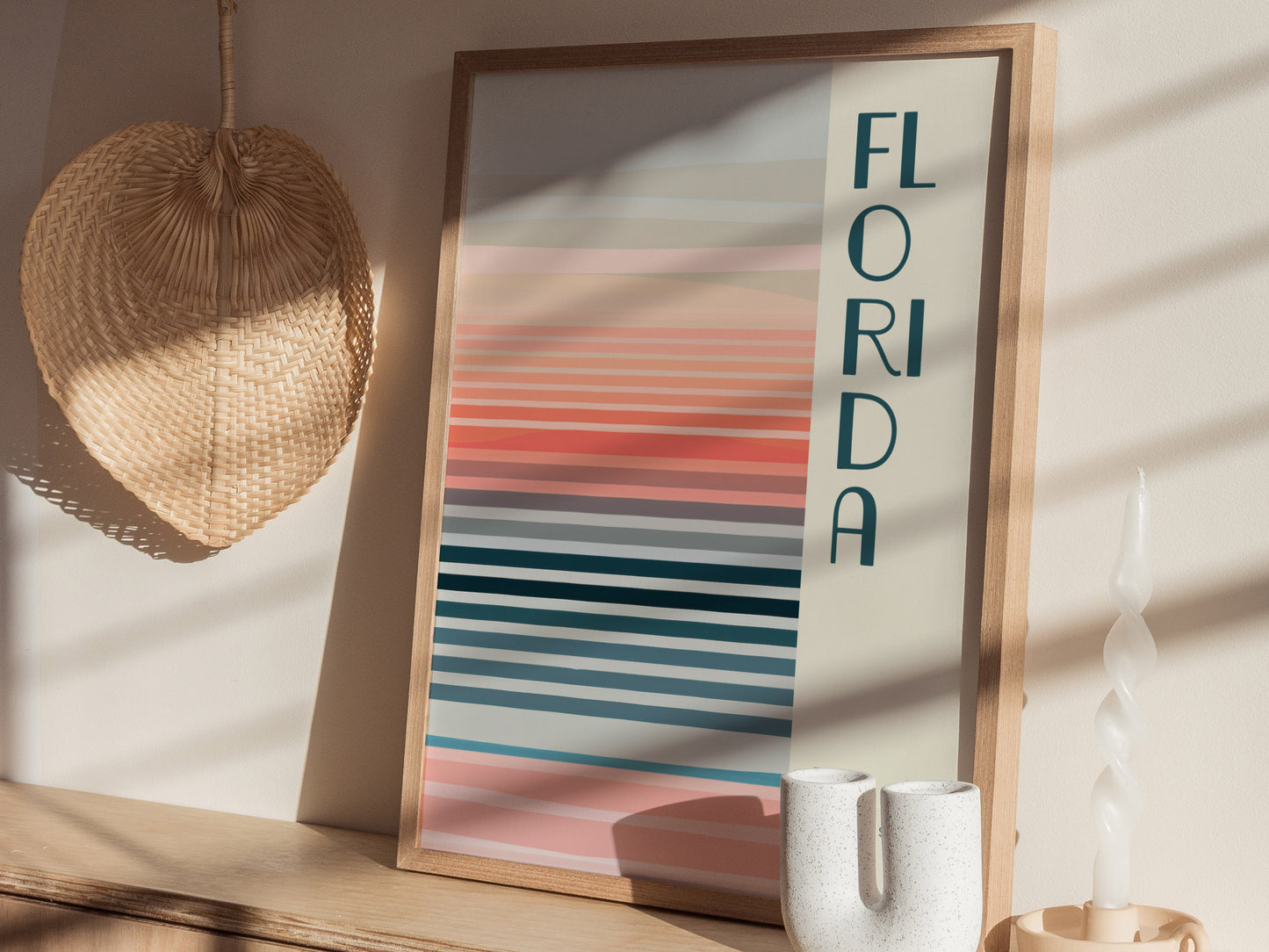 Framed poster with FLORIDA text and horizontal pastel stripes in pink, blue, and orange hues. Displayed on a wooden shelf with a woven fan, candle, and ceramic vase.