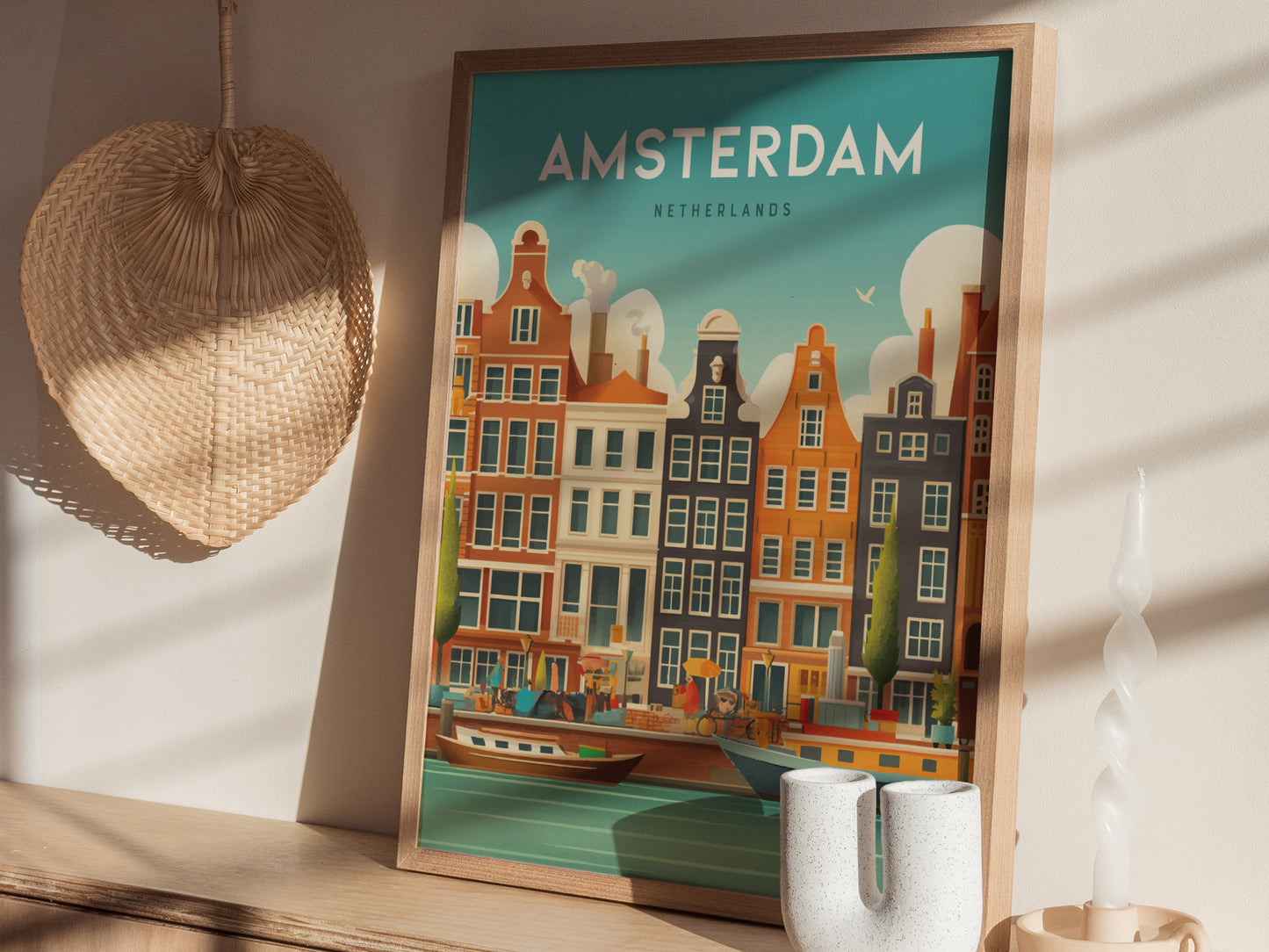 A framed Amsterdam poster depicting colorful canal houses is displayed on a shelf next to a woven fan and a white decorative candle. Sunlight casts shadows on the wall.