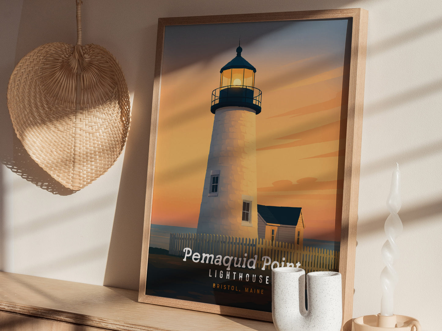 A framed art print of the Pemaquid Point Lighthouse in Bristol, Maine, is displayed on a wall. The artwork features a serene sunset with warm hues illuminating the lighthouse and surrounding landscape, creating a tranquil coastal scene.