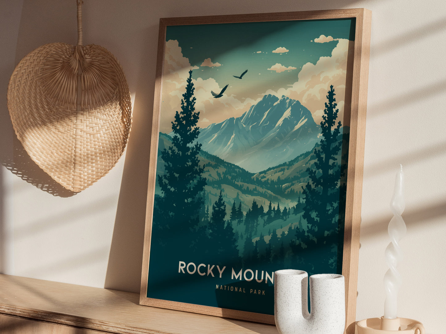 A framed Rocky Mountain National Park poster with mountains and trees stands on a table next to a woven basket, decorative vase, and twisted candle in soft lighting.