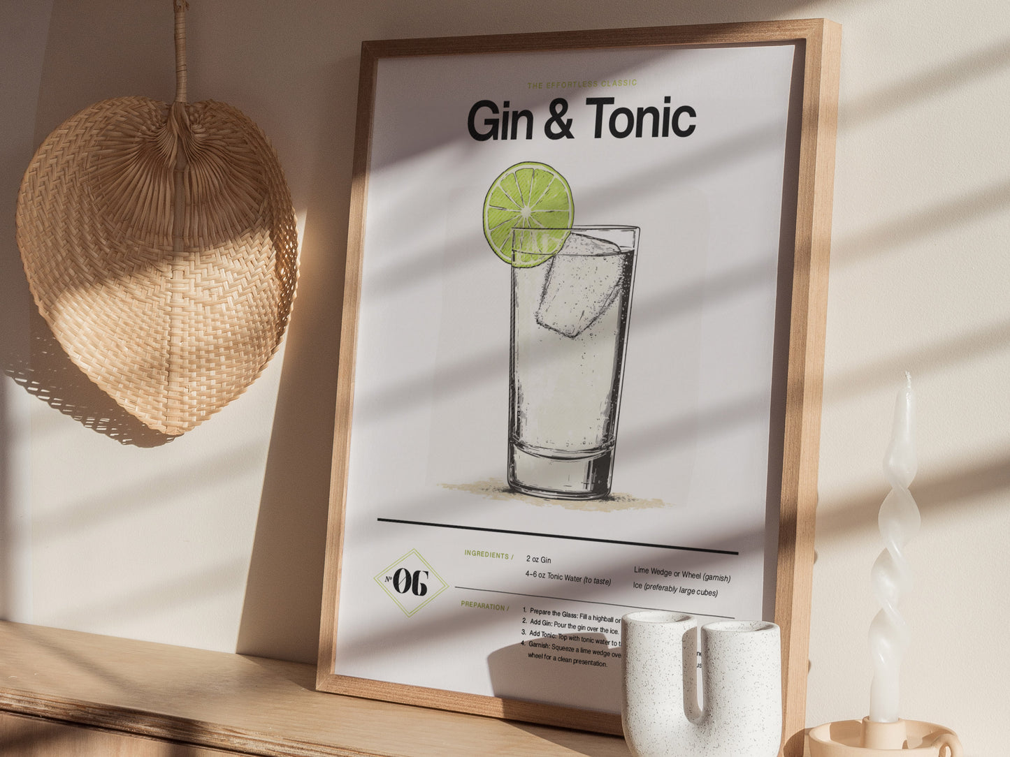 A framed gin and tonic cocktail poster with a minimalist design is displayed on a shelf next to a woven wall fan and a twisted white candle.