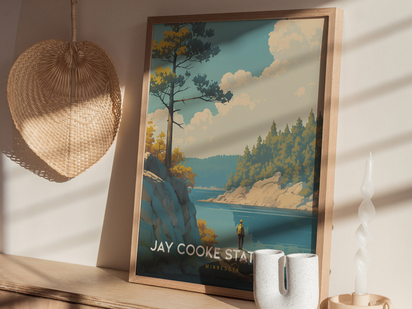 A framed poster of Jay Cooke State Park is displayed on a wooden surface, featuring a scenic river and forest. Sunlight casts shadows across a woven basket and a candle.
