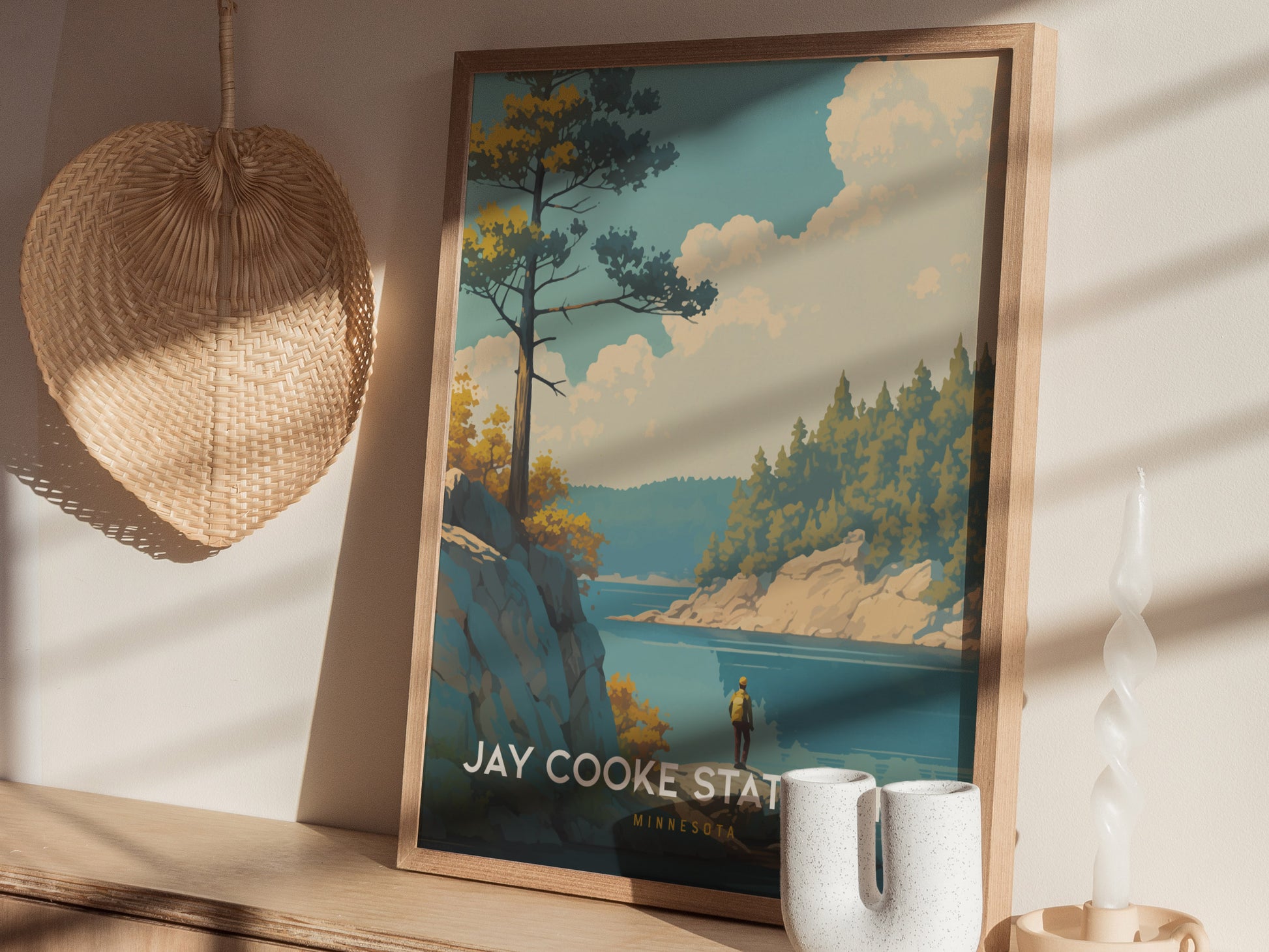 A framed poster of Jay Cooke State Park is displayed on a wooden surface, featuring a scenic river and forest. Sunlight casts shadows across a woven basket and a candle.