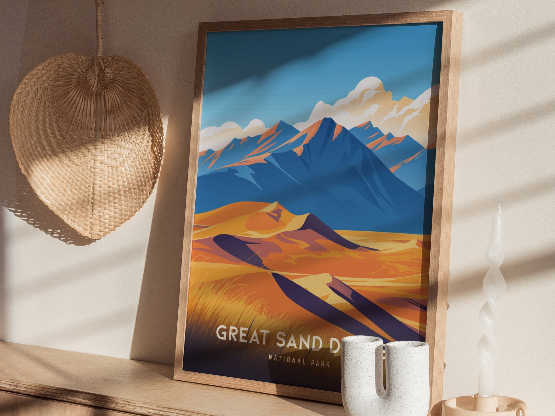 A framed poster of Great Sand Dunes National Park with colorful mountains and dunes is displayed on a wooden surface, next to a straw hat and a twisted white candle.