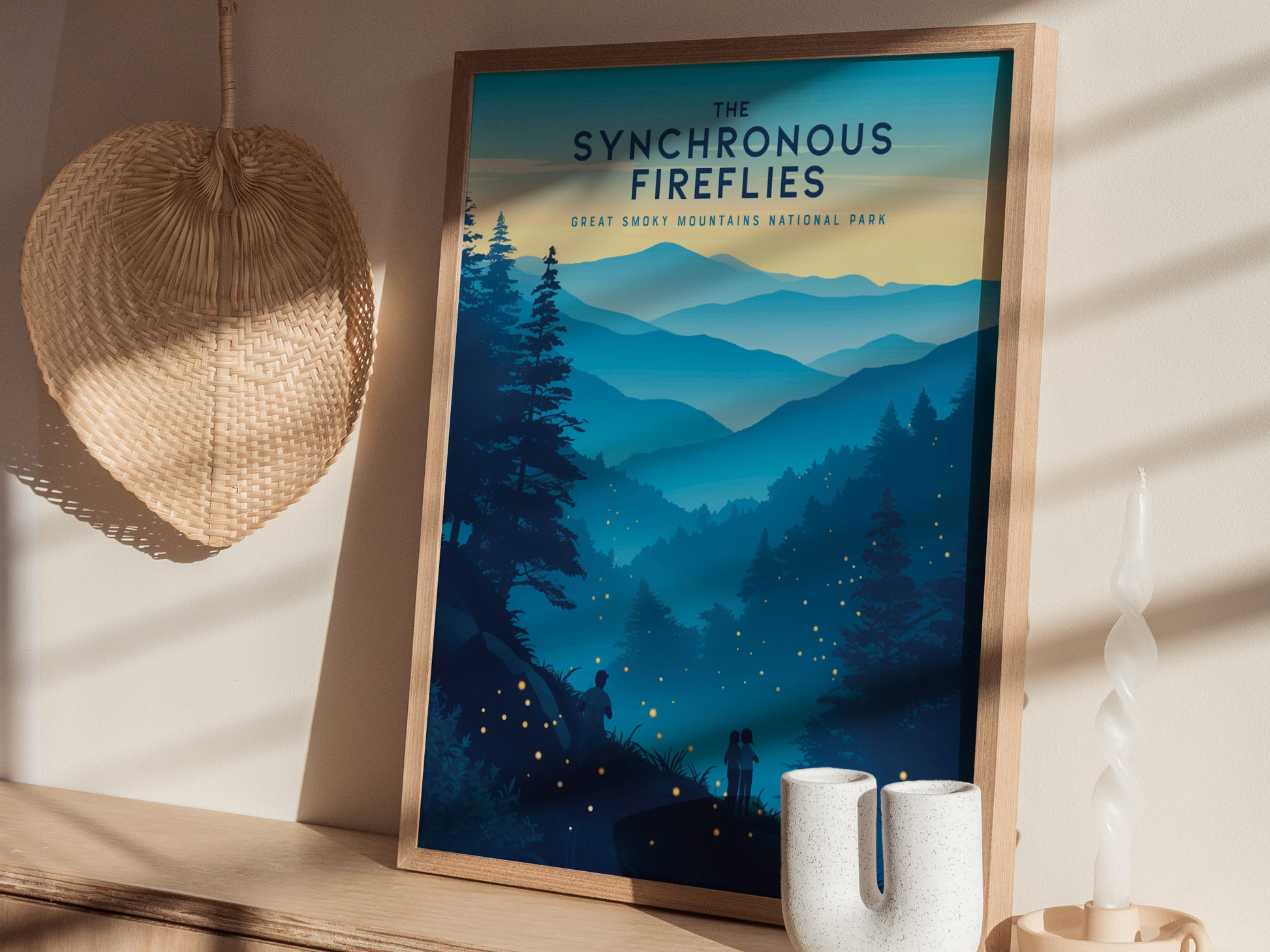 A framed poster titled The Synchronous Fireflies depicts a serene landscape of the Great Smoky Mountains at dusk, with fireflies illuminating the scene. The poster is displayed on a wooden shelf beside a woven wall decor and a white candle.