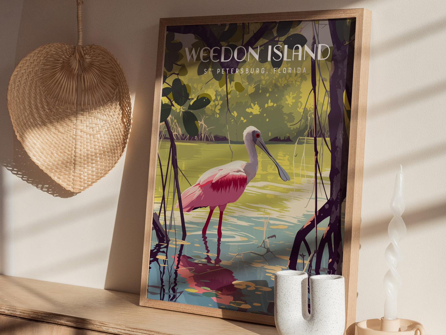 A framed poster of a pink Roseate Spoonbill wading through mangrove waters is displayed on a wooden shelf, accompanied by a woven fan and decorative candle.