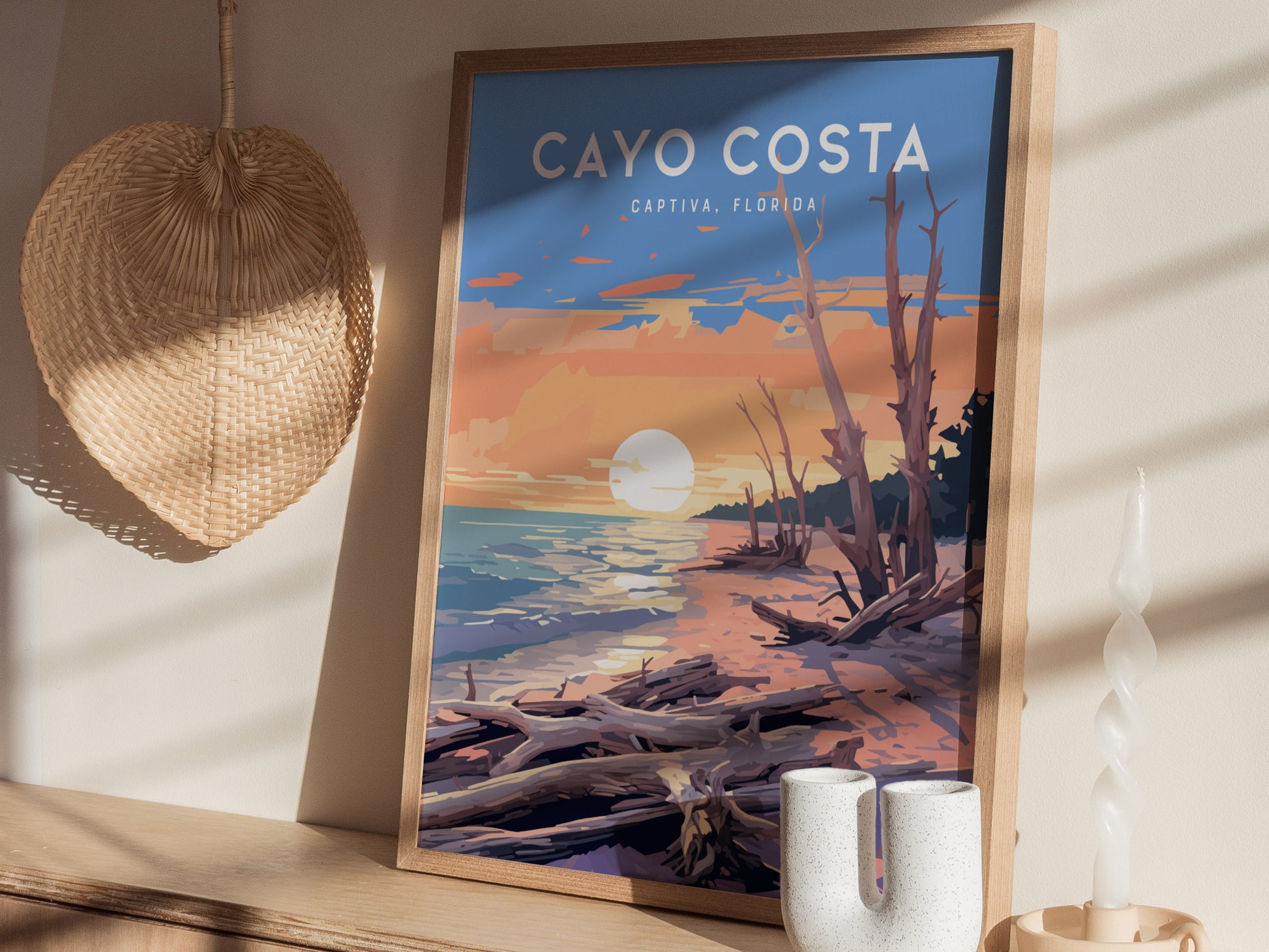 A framed Cayo Costa State Park poster depicting a vibrant sunset over a shoreline with driftwood. The scene is set on a wooden shelf, accompanied by a woven wall decoration.