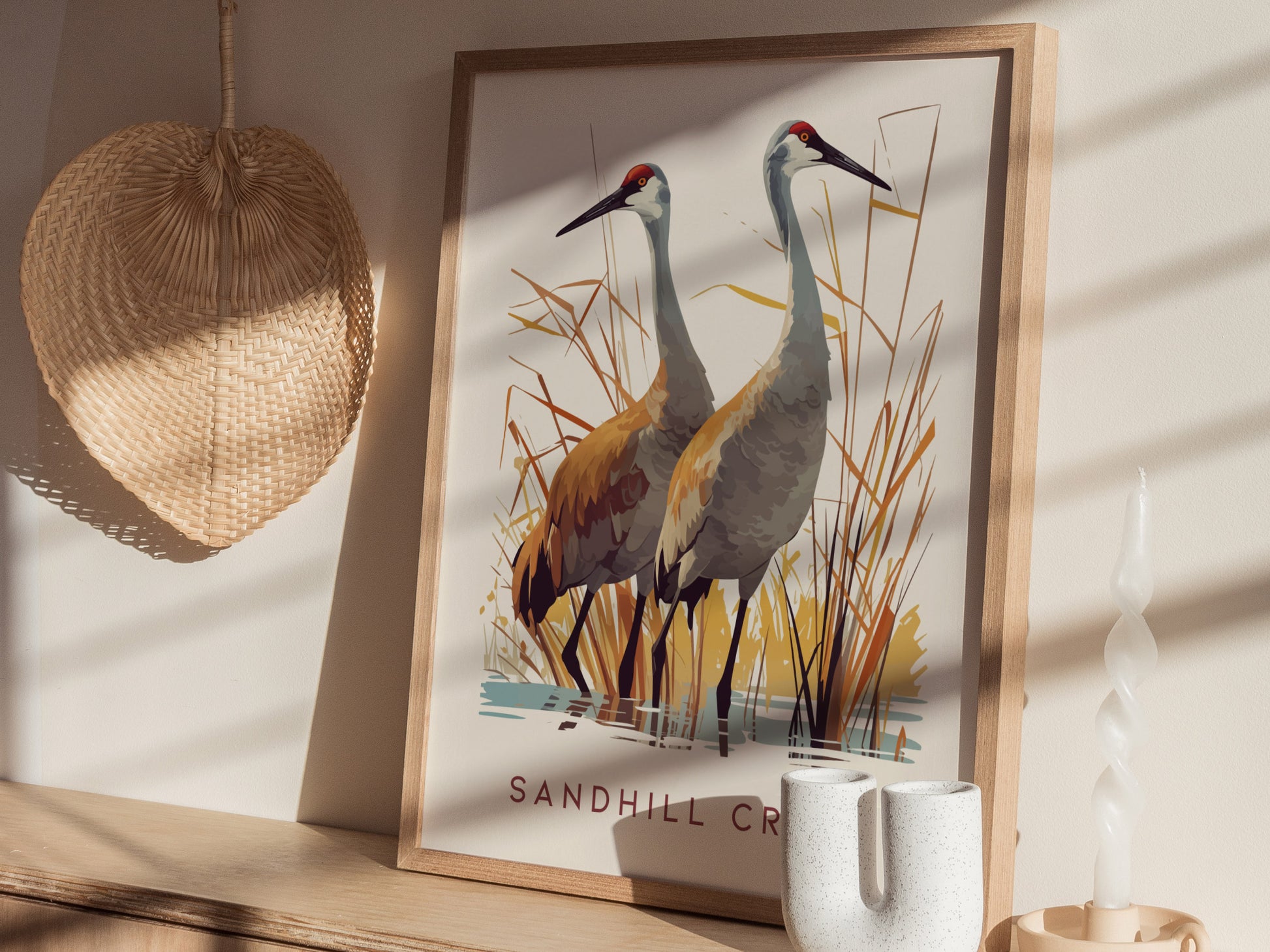The image shows a framed Sandhill Cranes poster on a shelf, surrounded by a woven fan, a twisted white candle, and a decorative vase. Sunlight casts shadows on the wall.